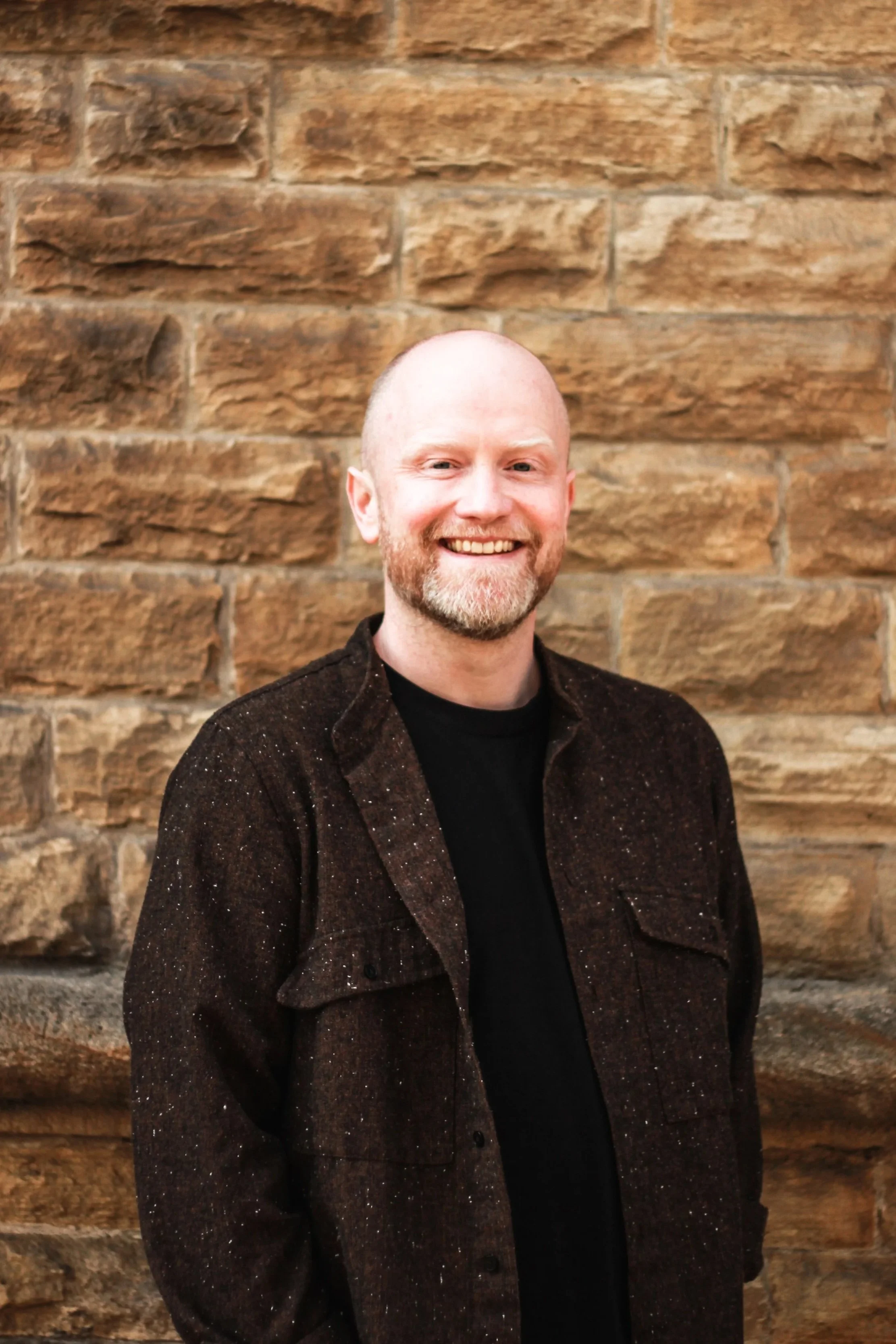 A smiling man with a beard and mustache standing in front of a brick wall.