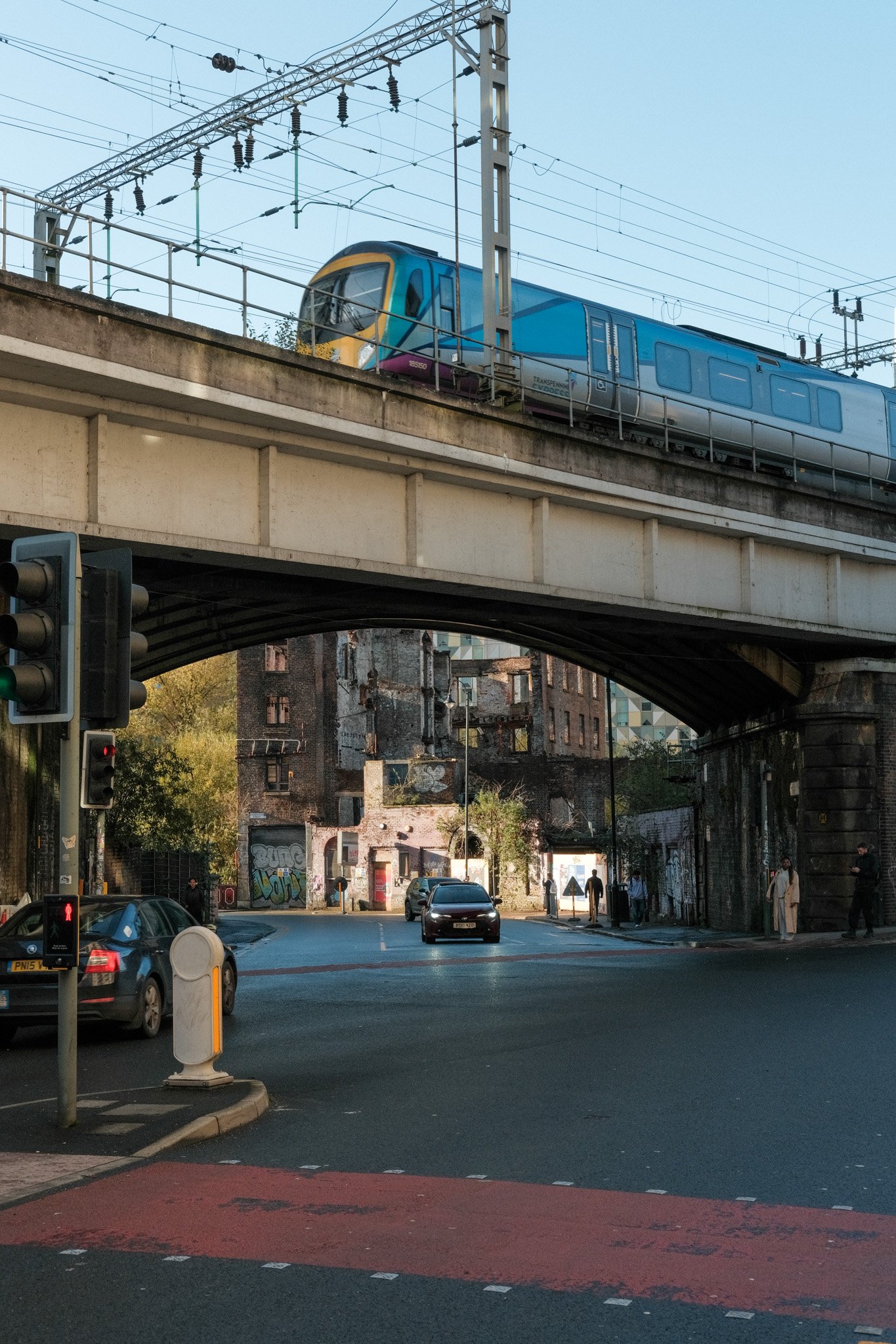 A blue train on an elevated track passing above a city street with cars, pedestrians, and graffiti-covered buildings in the background. Traffic lights and street signs are visible.