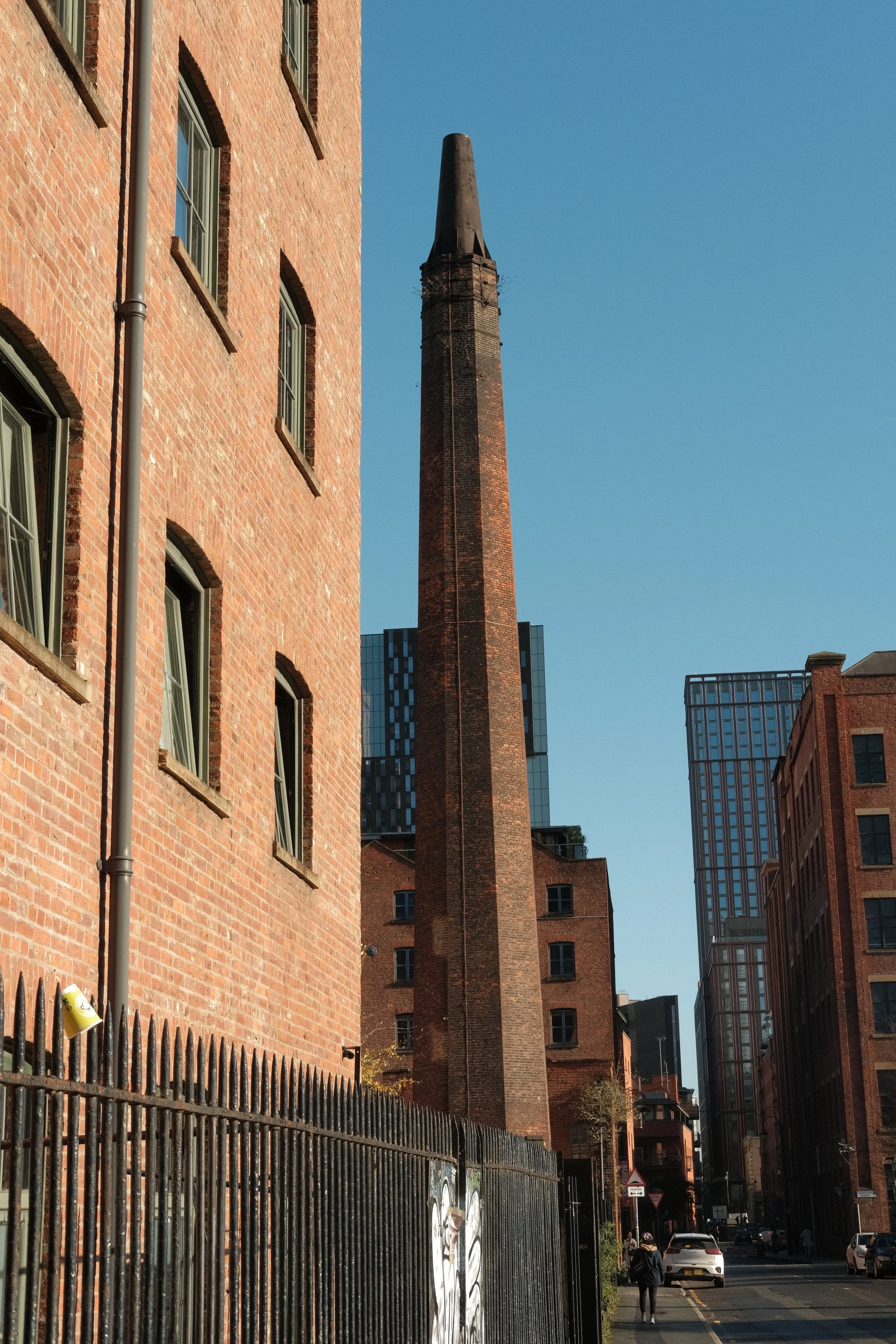 City street scene with brick buildings, a tall smokestack, and modern glass high-rise buildings in the background under a clear blue sky.