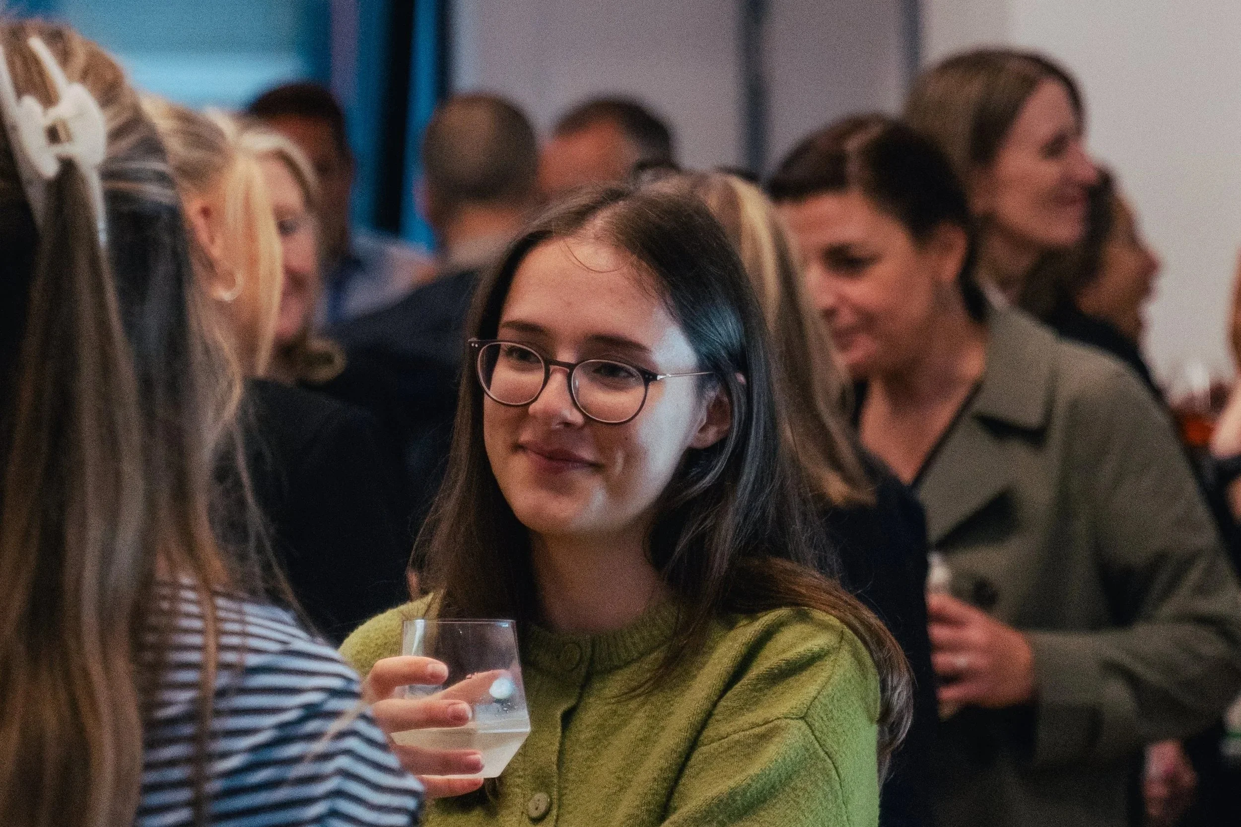 A woman with long black hair and glasses wearing a green sweater, holding a glass, talking to another woman with blond hair and striped shirt, at a social gathering with multiple people in the background.