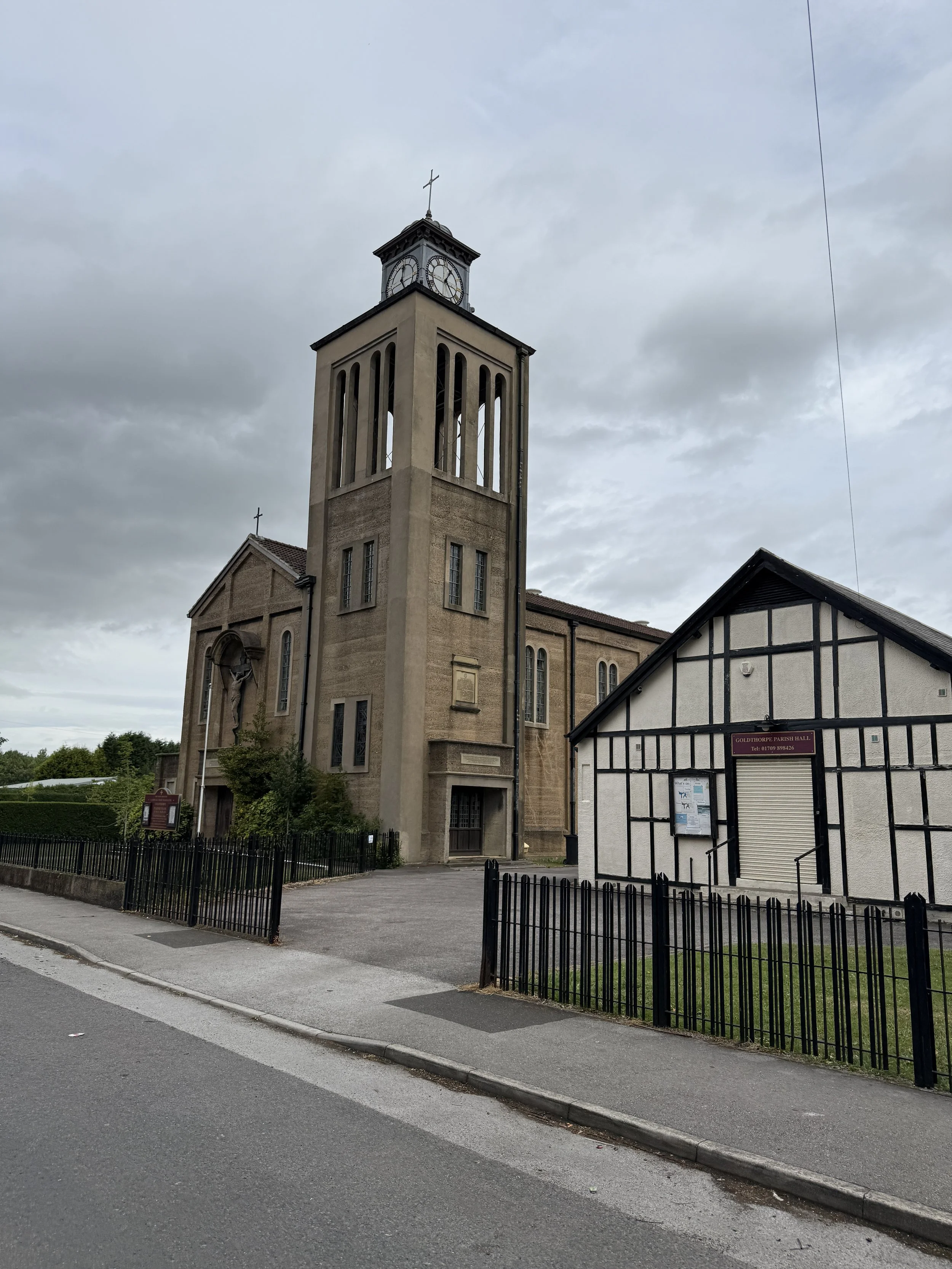 A church with a tall clock tower and a cross at the top, next to a smaller building with a sign that reads "GOLDHOPPE PARISH HALL". The surroundings include a paved street, a fence, and some trees, under a cloudy sky.