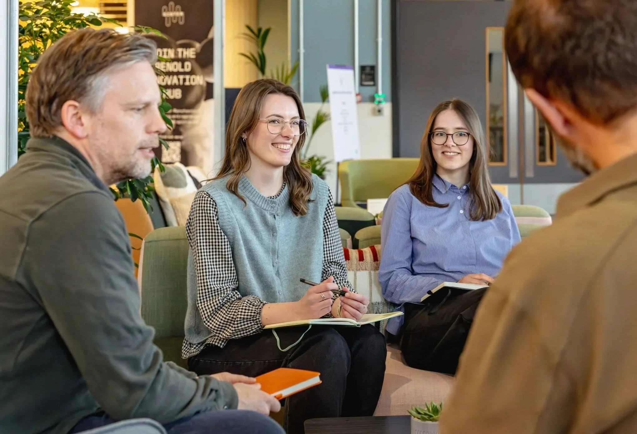 A group of four young adults sitting in a circle in a modern indoor space, engaging in conversation. Two women are smiling, one even appears to be speaking, while a man holds a notebook and a pen in the foreground. The setting features plants, a dark wall with framed decor, and informational signs.