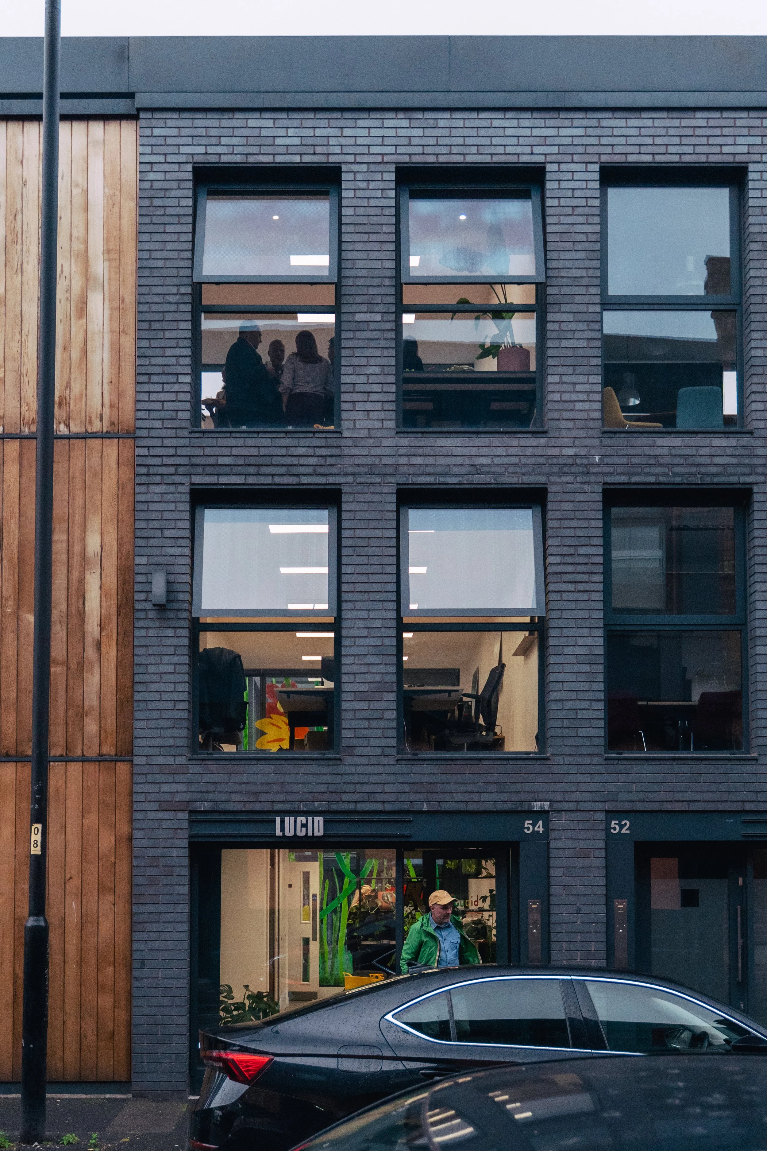 Urban street scene with a modern building featuring a mix of brick and wood exterior, and large glass windows. People visible through the windows inside the building, and a parked car in the foreground.