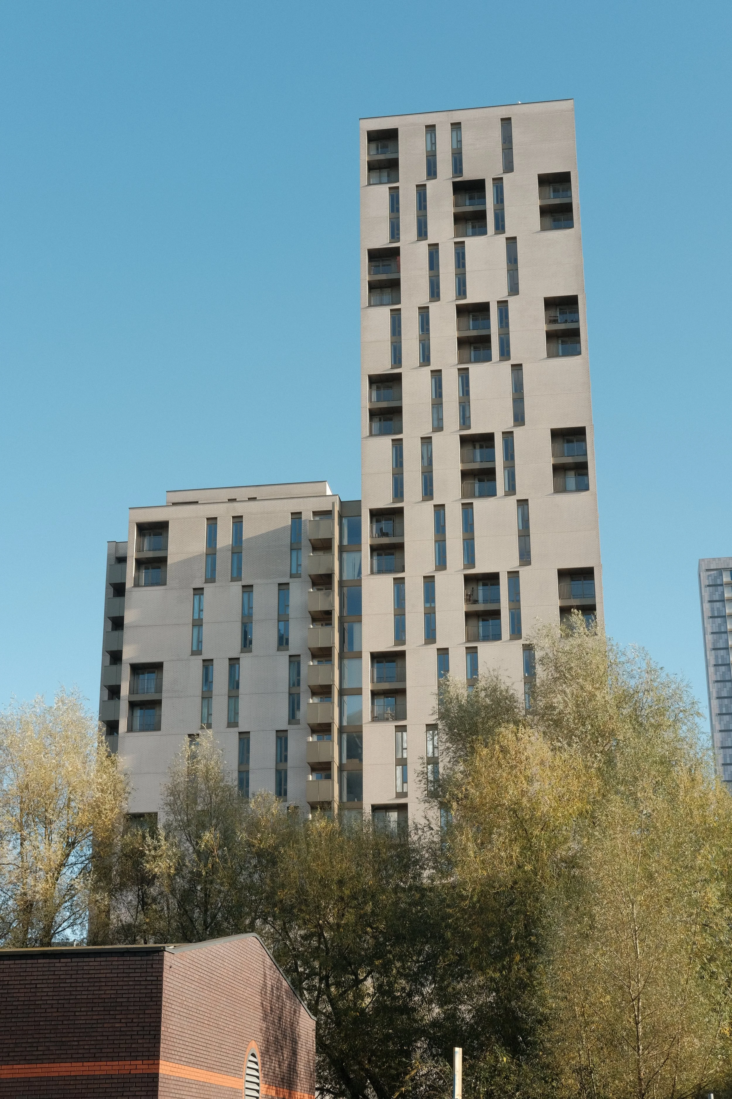 Tall modern residential building with unique geometric window patterns, set against a clear blue sky, with trees at the base.