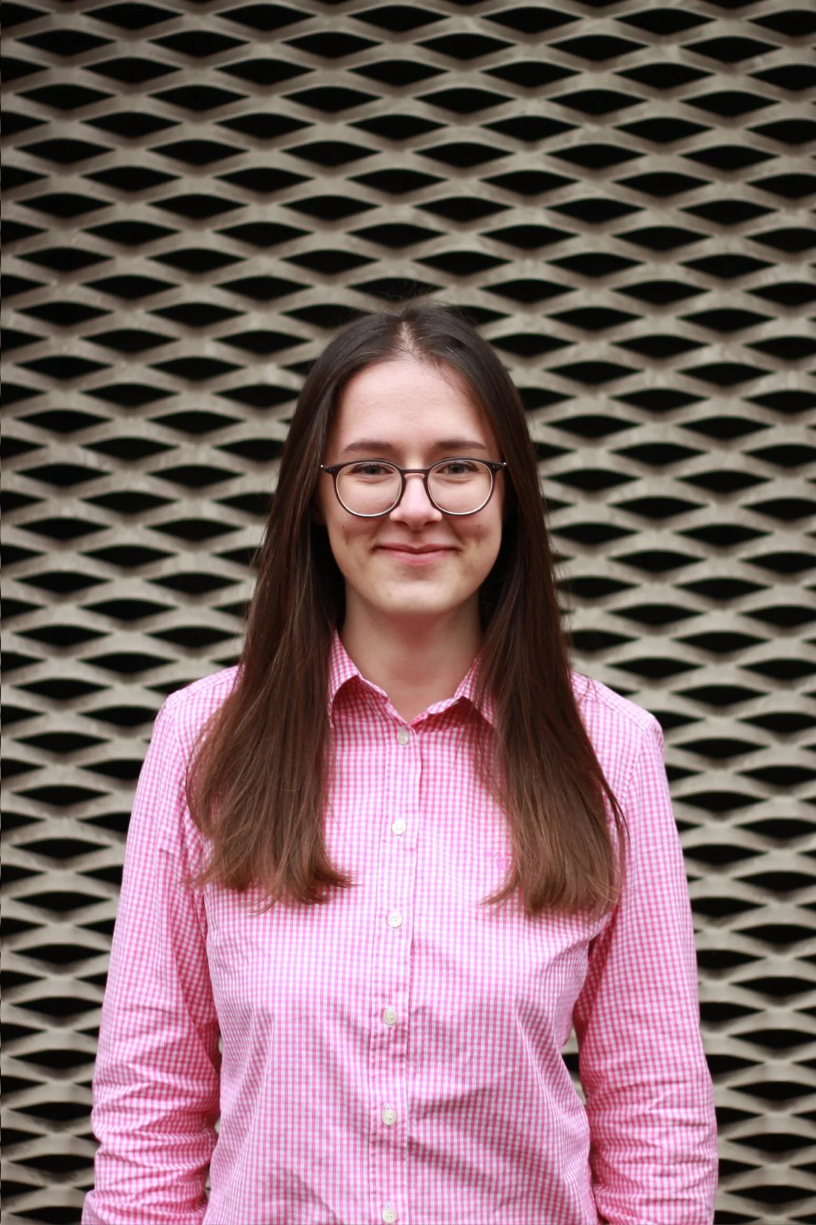 A young woman with long brown hair and glasses, wearing a pink checkered shirt, stands in front of a black and white geometric patterned wall.