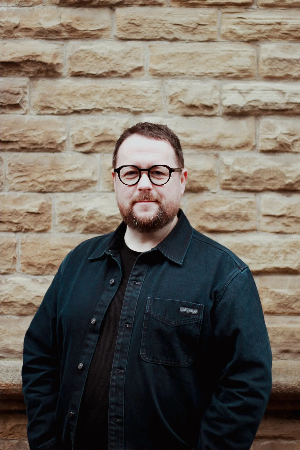 A man with glasses and a beard standing in front of a brick wall, wearing a black denim jacket.
