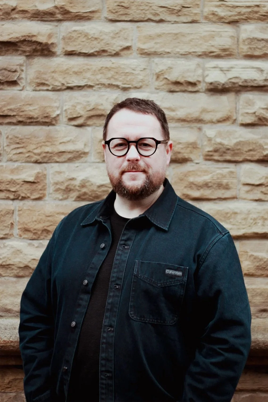 A man with glasses and a beard wearing a dark denim shirt standing in front of a beige brick wall.