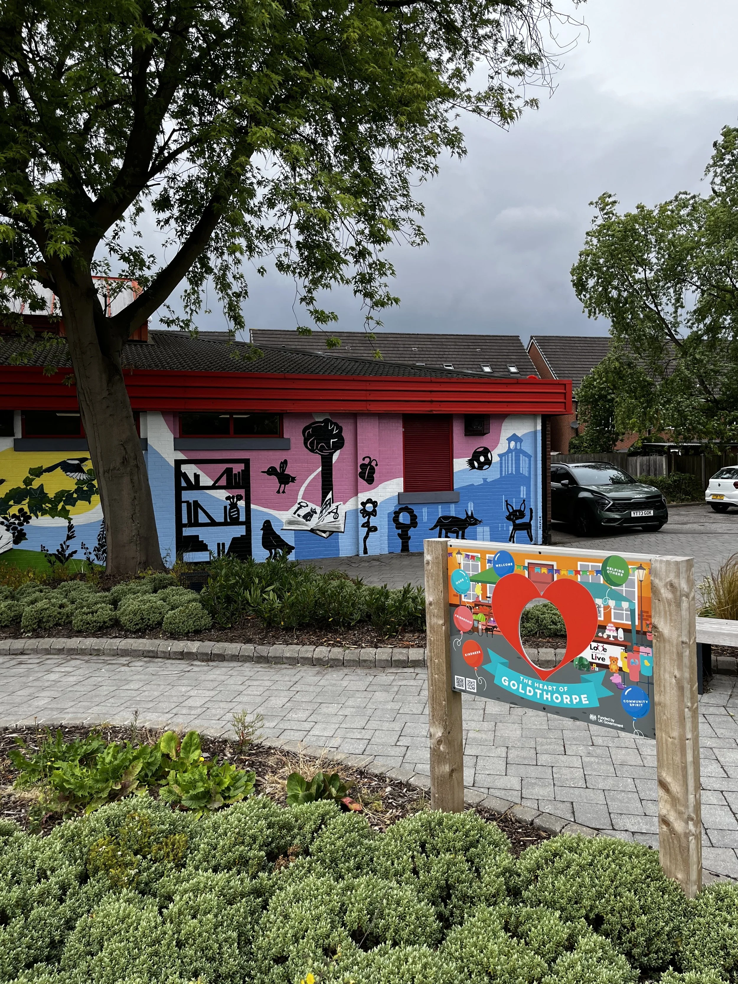 A mural painted on a building wall featuring black silhouette animals, trees, and a book, with a cloud landscape background. In the foreground, a colorful sign with balloons, a heart symbol, and the text 'The heart of Goldthorpe' is visible. There are parked cars and trees along a sidewalk with greenery and flowers.