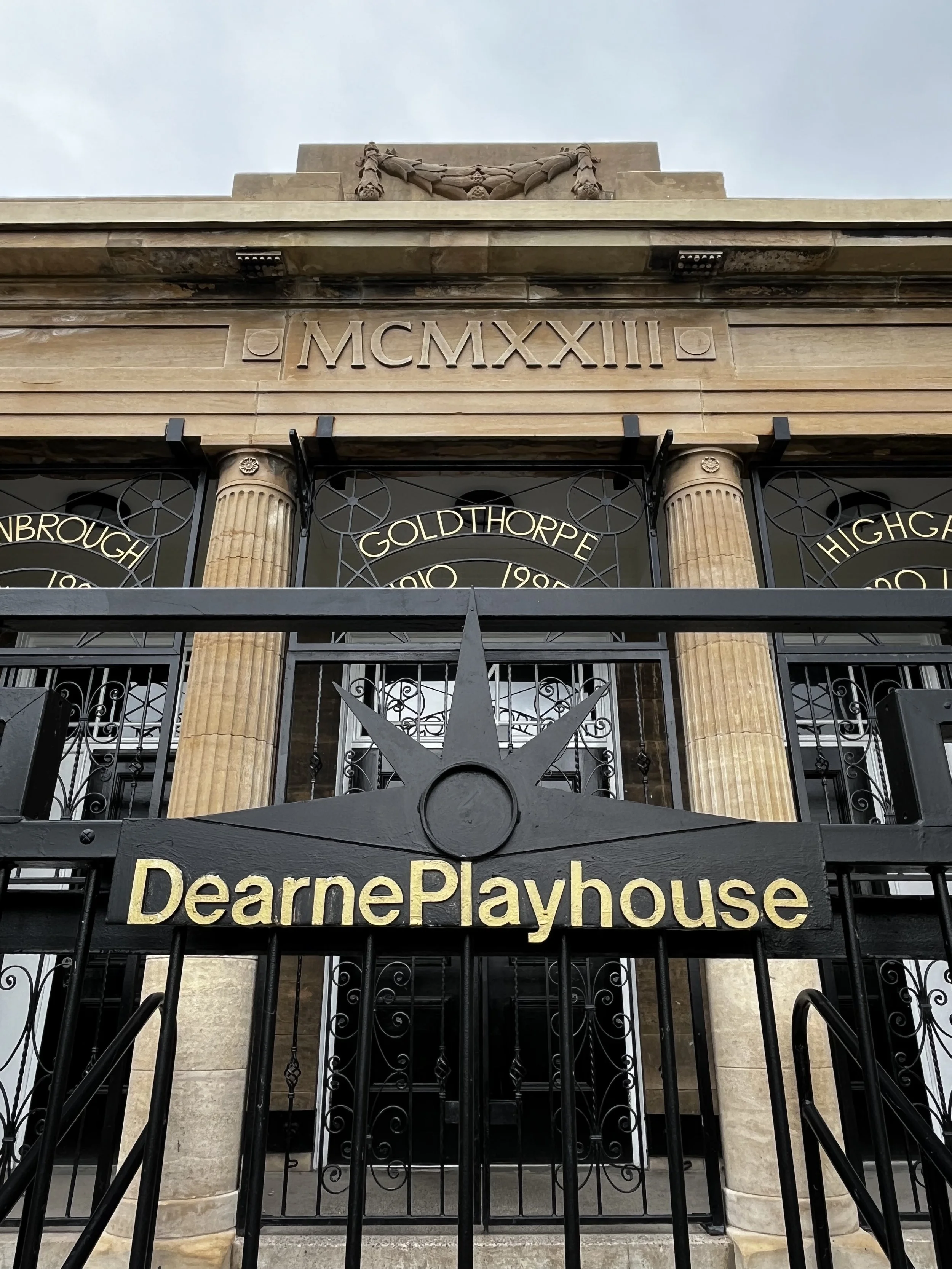 Front view of Dearne Playhouse, a building with classical columns and a wrought iron gate in front, with the name 'Dearne Playhouse' in gold letters on a black background.