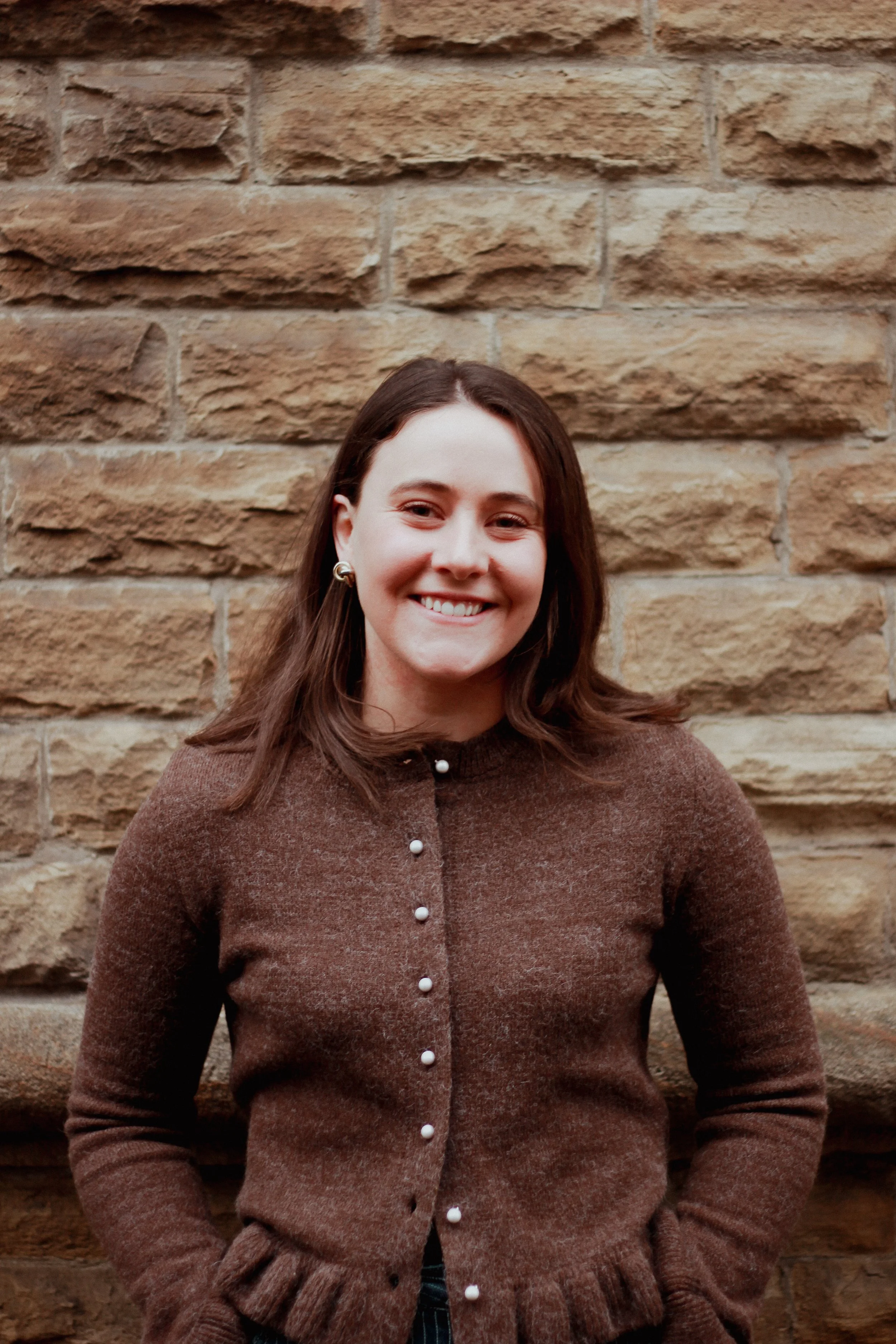 A woman smiling and standing in front of a brick wall, wearing a brown cardigan with pearl buttons.