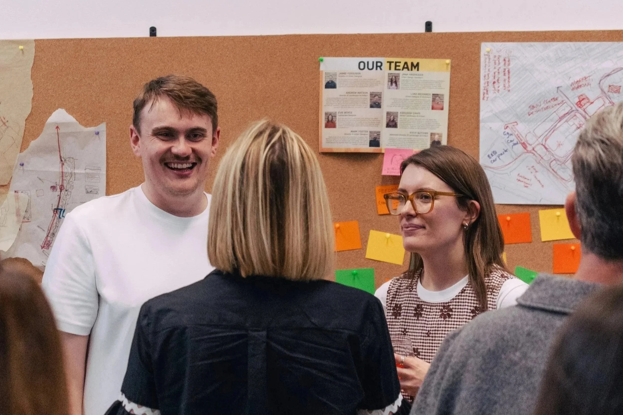 A group of people in a discussion or meeting, with a focus on a young man smiling at the camera. Others are engaged in conversation, and posters with maps and team information are visible on the corkboard in the background.