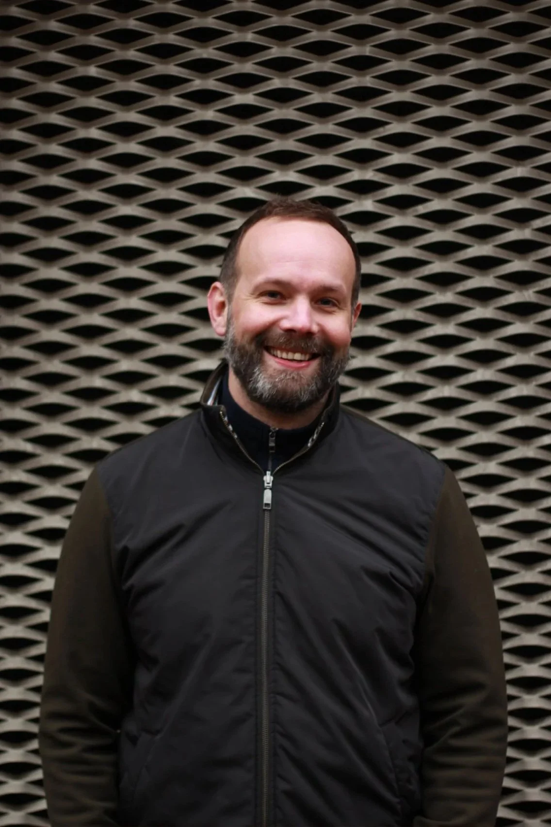 A smiling man with a beard and short hair standing in front of a textured, diamond-shaped wall.
