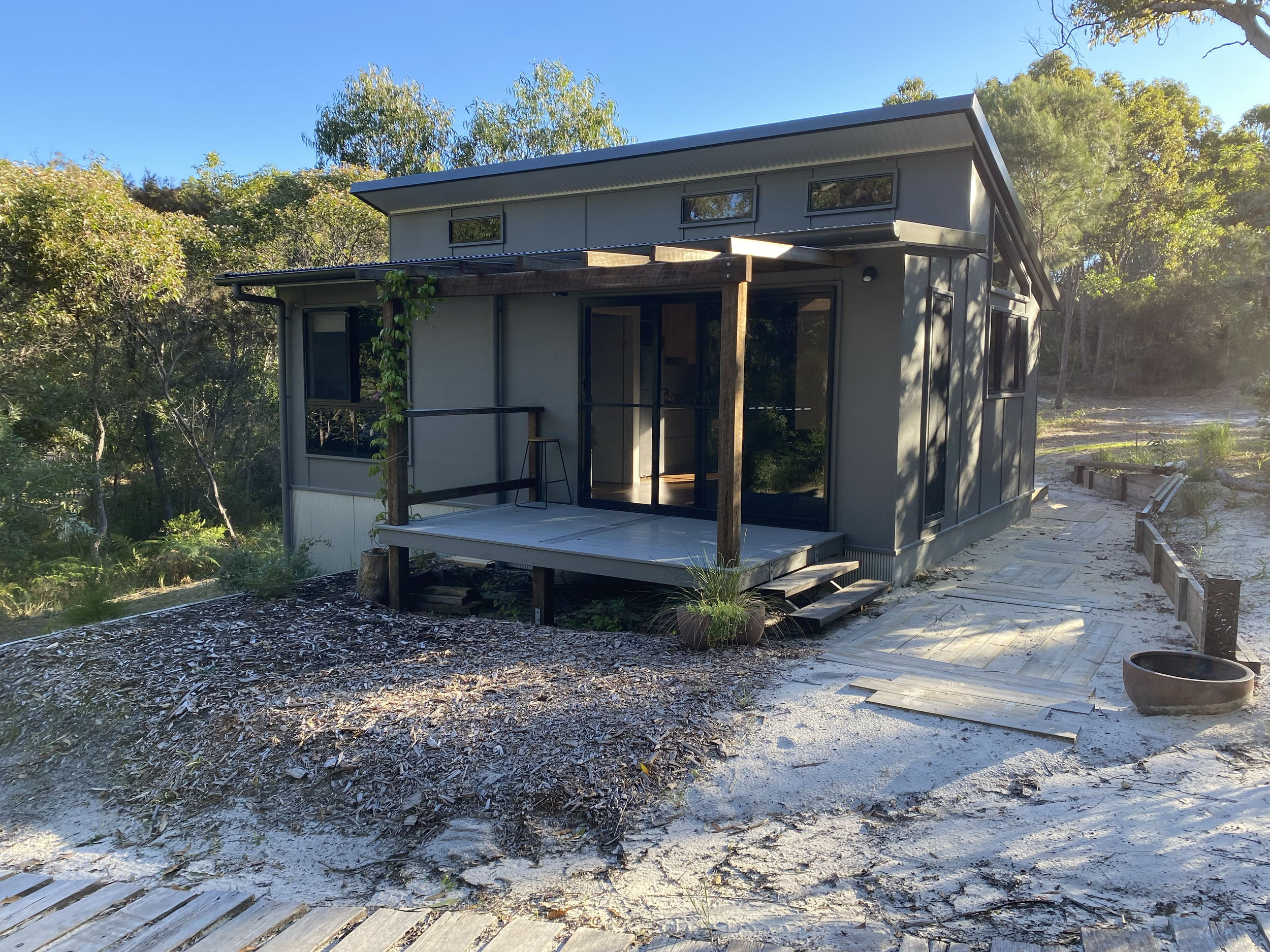 A small modern house with a gray exterior and a sloped metal roof, built on a sandy landscape with trees in the background, featuring a front porch with a wooden deck and steps. Orchid Beach accommodation K'gari Fraser Island