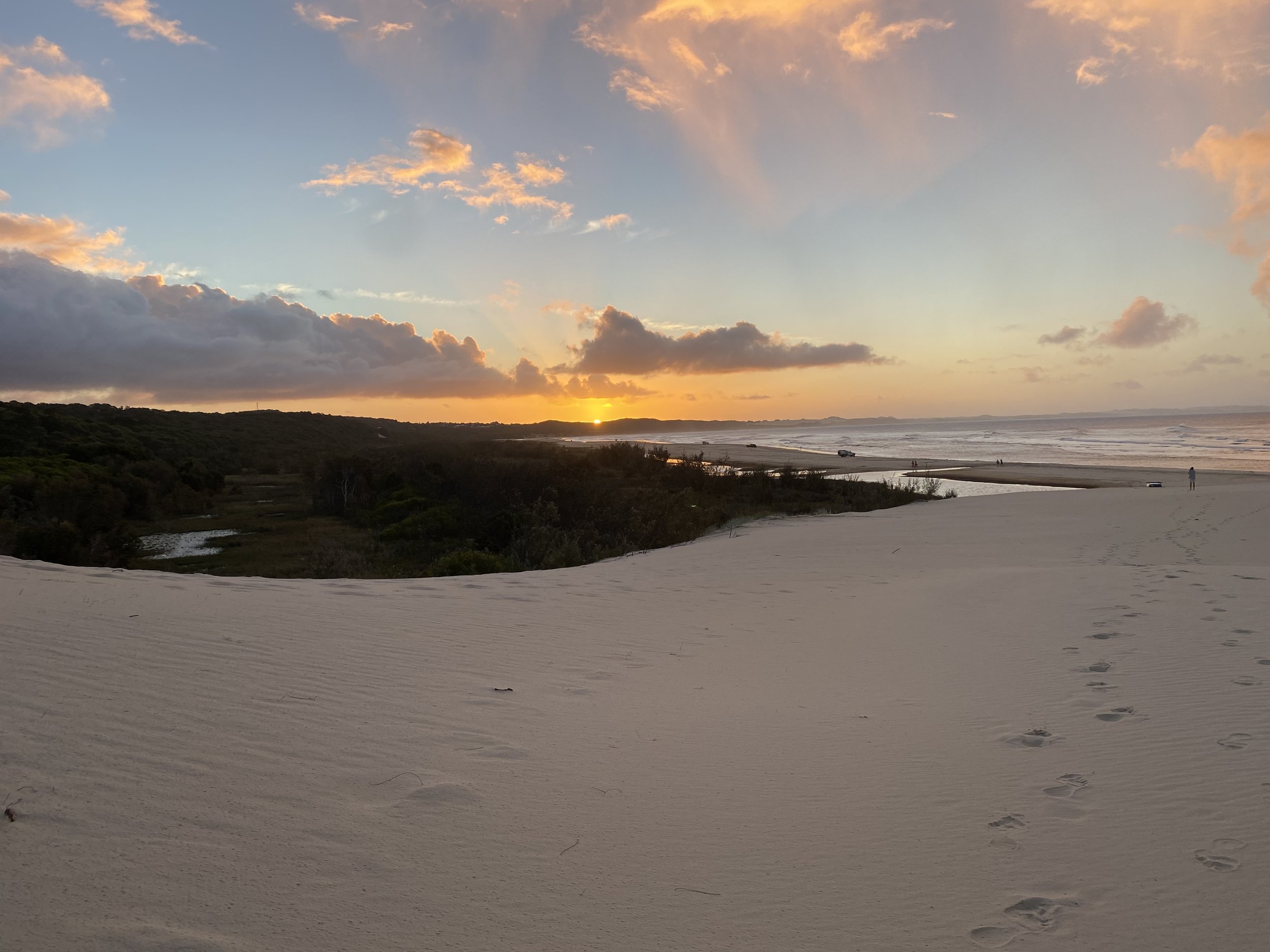 A beach at sunset with footprints in the sand, a few people in the distance, and the ocean waves, sky with clouds and the sun near the horizon.