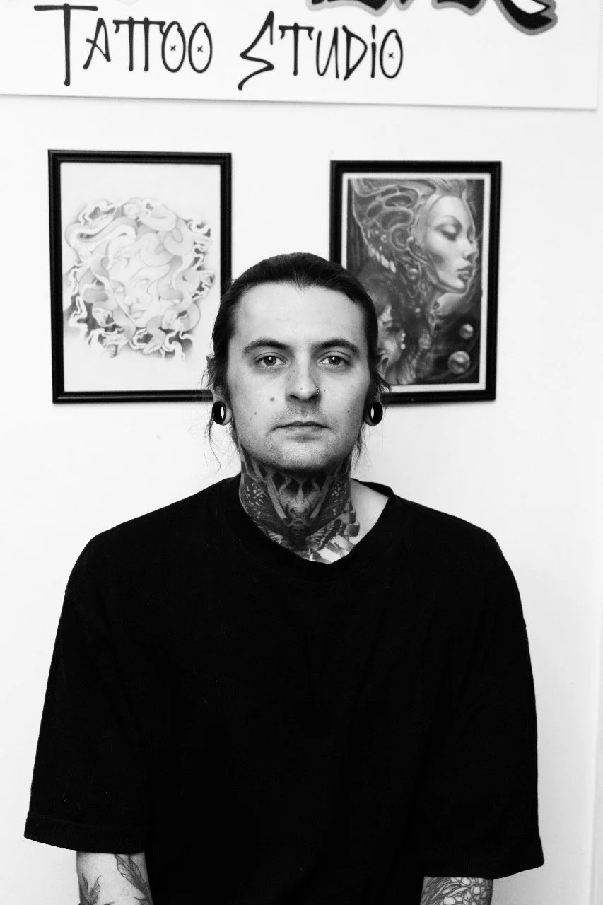 Black and white photo of a man with tattoos and piercings sitting in front of tattoo artwork, with a sign reading 'Tattoo Studio' above him.