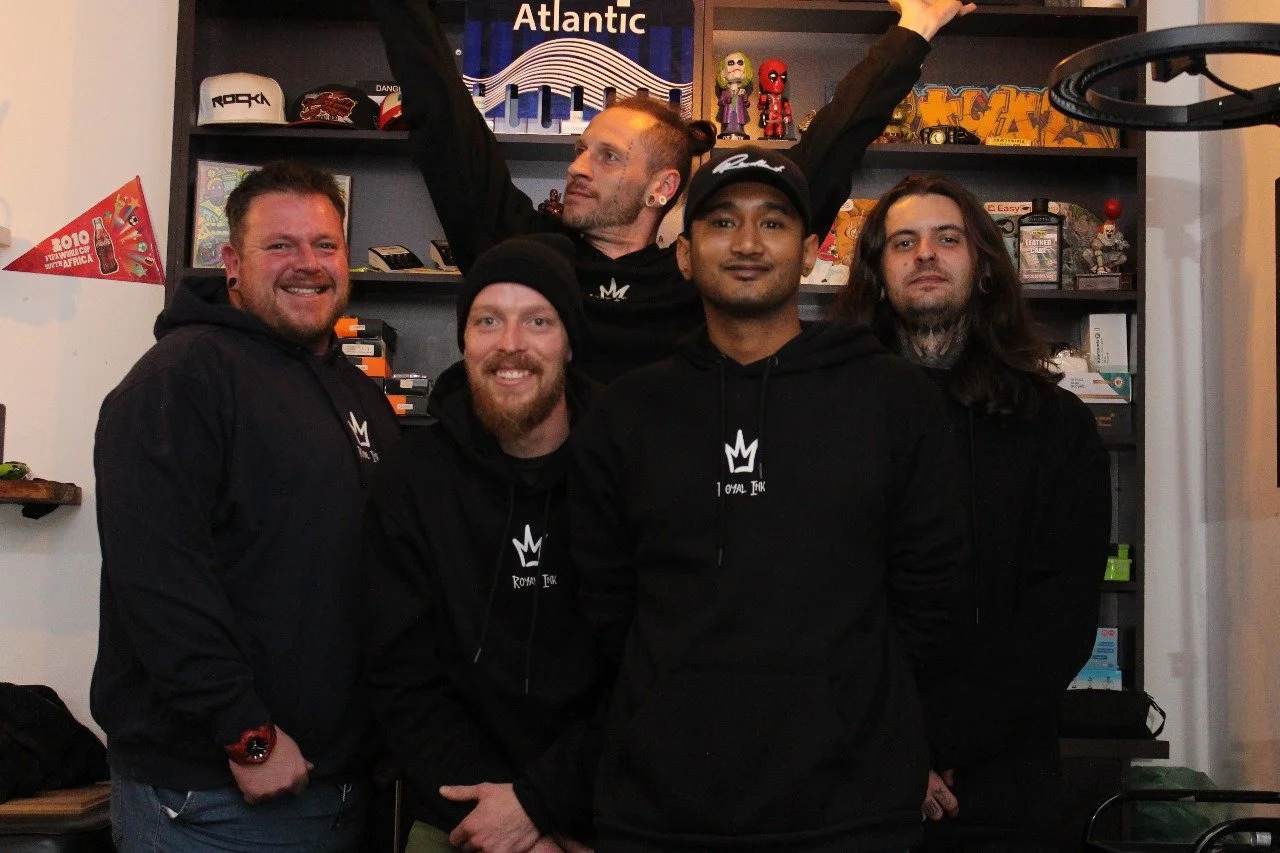 Group of five men posing indoors, some wearing black hoodies with a white crown logo, shelves with collectibles and memorabilia in the background.