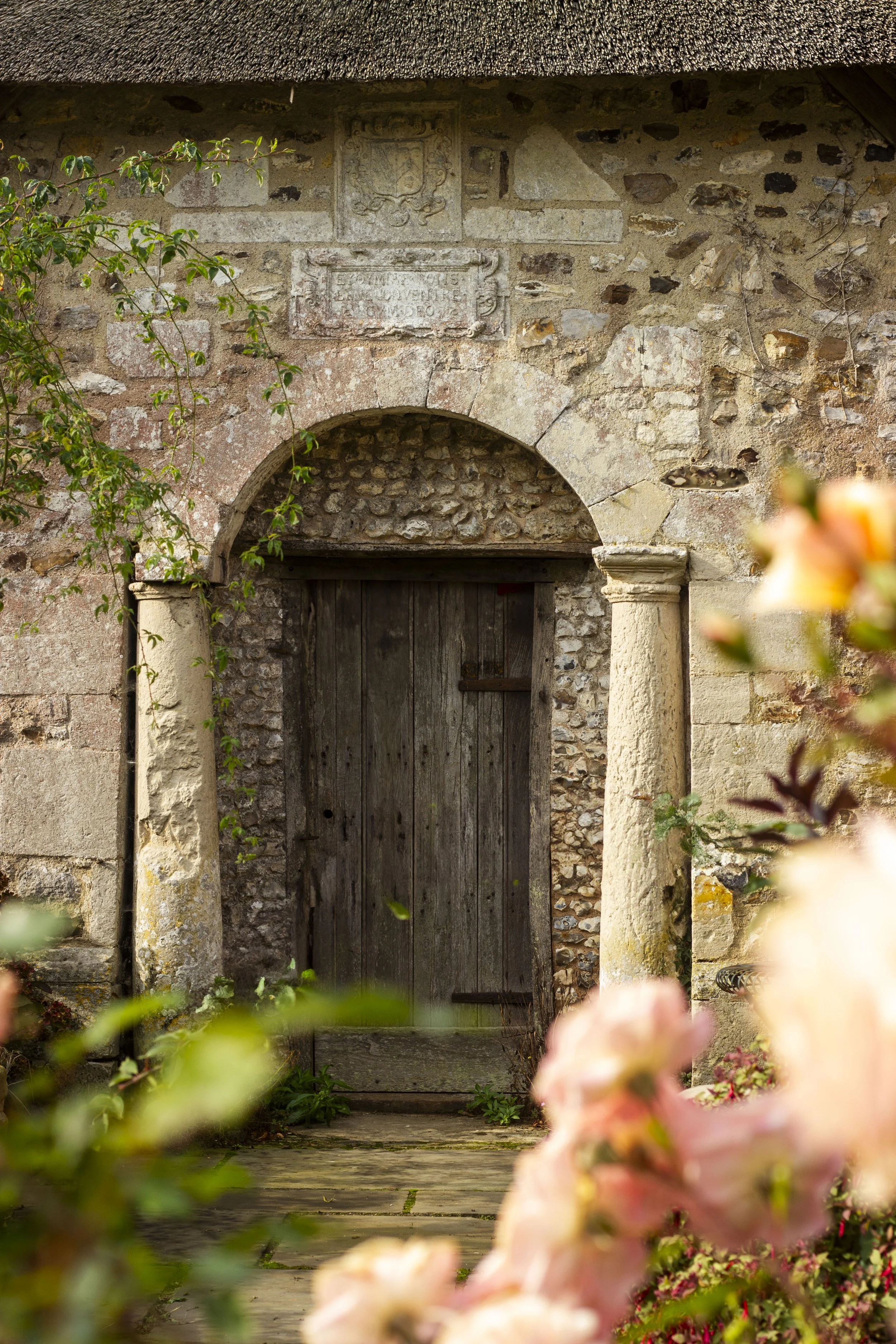Old stone and wood doorway with an arch, framed by columns, and surrounded by greenery and flowers.