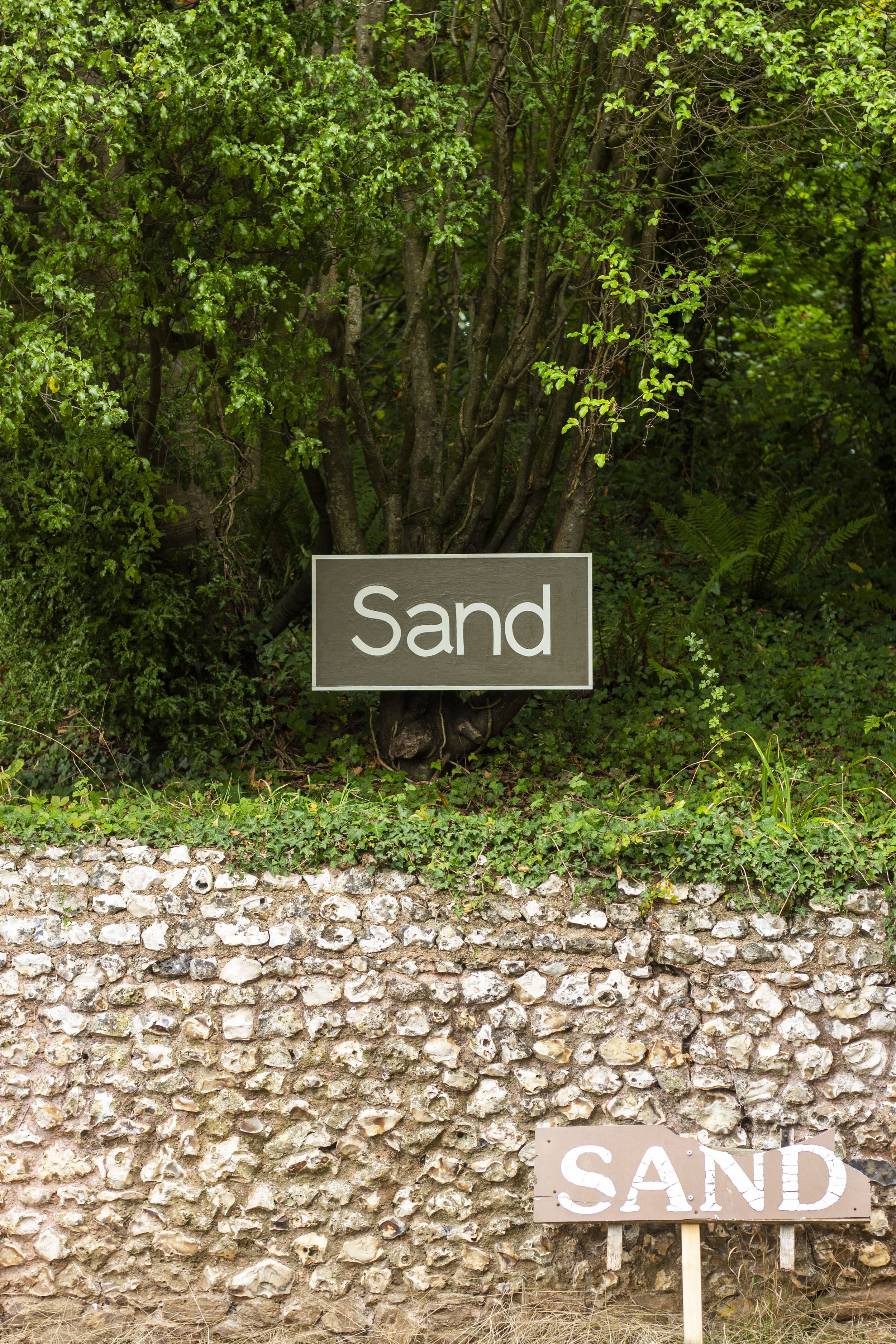A wooded area with green foliage and a stone wall in the foreground. There are two signs: a dark brown sign with white text that reads 'Sand' attached to the tree and placed on the ground, and a partially visible sign with the word 'SAND' in white capital letters on a light background in front of the stone wall.