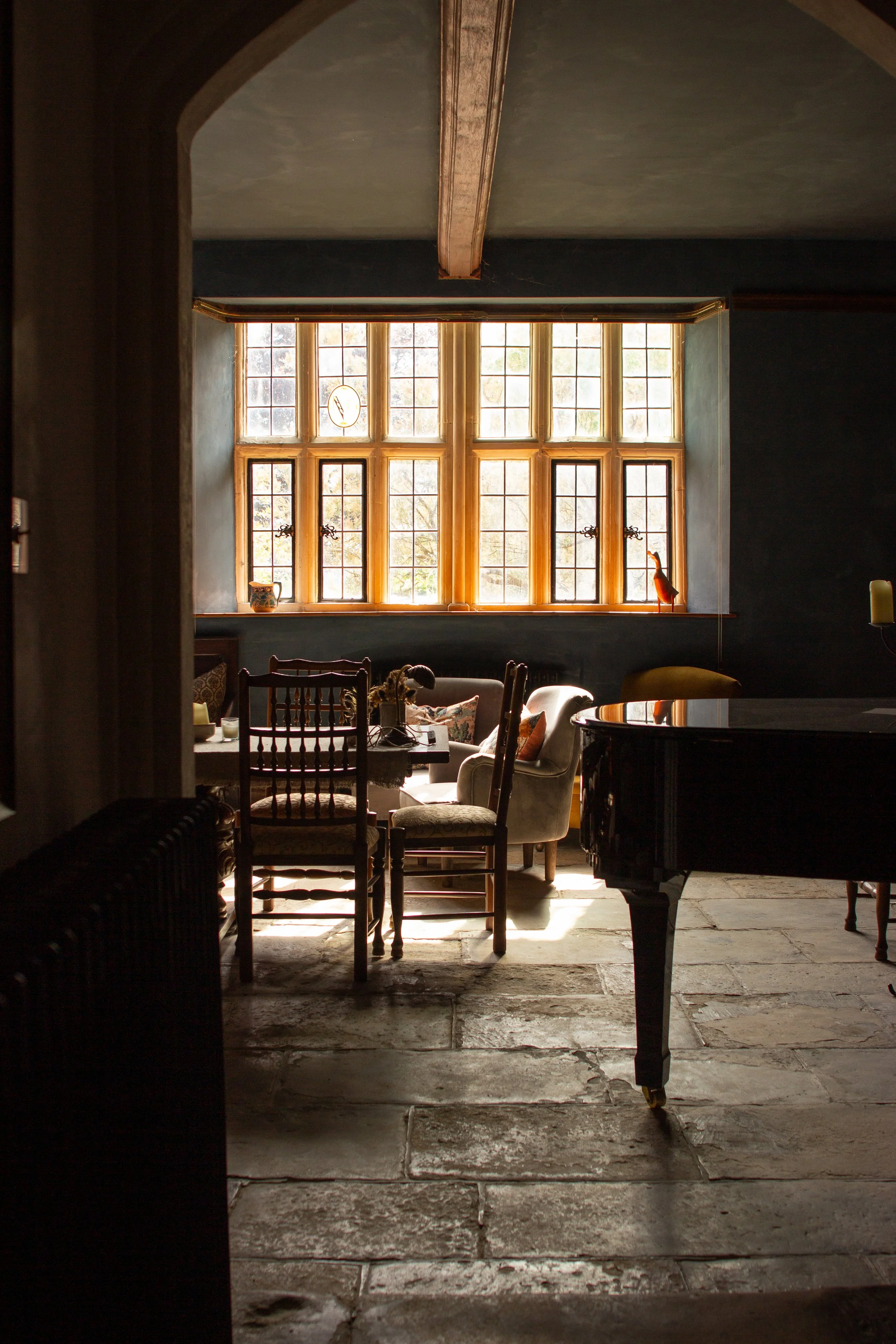 View through an arched doorway into a dimly lit room with a large window. The room has vintage furniture including a table, chairs, armchairs, and a grand piano. The window is illuminated by sunlight, casting shadows inside the room.