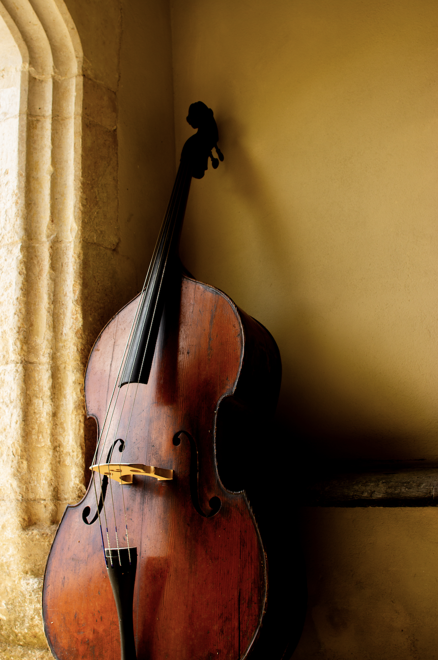 An old wooden double bass leaning against a wall with a window and beige curtain nearby, inside a room with warm lighting.