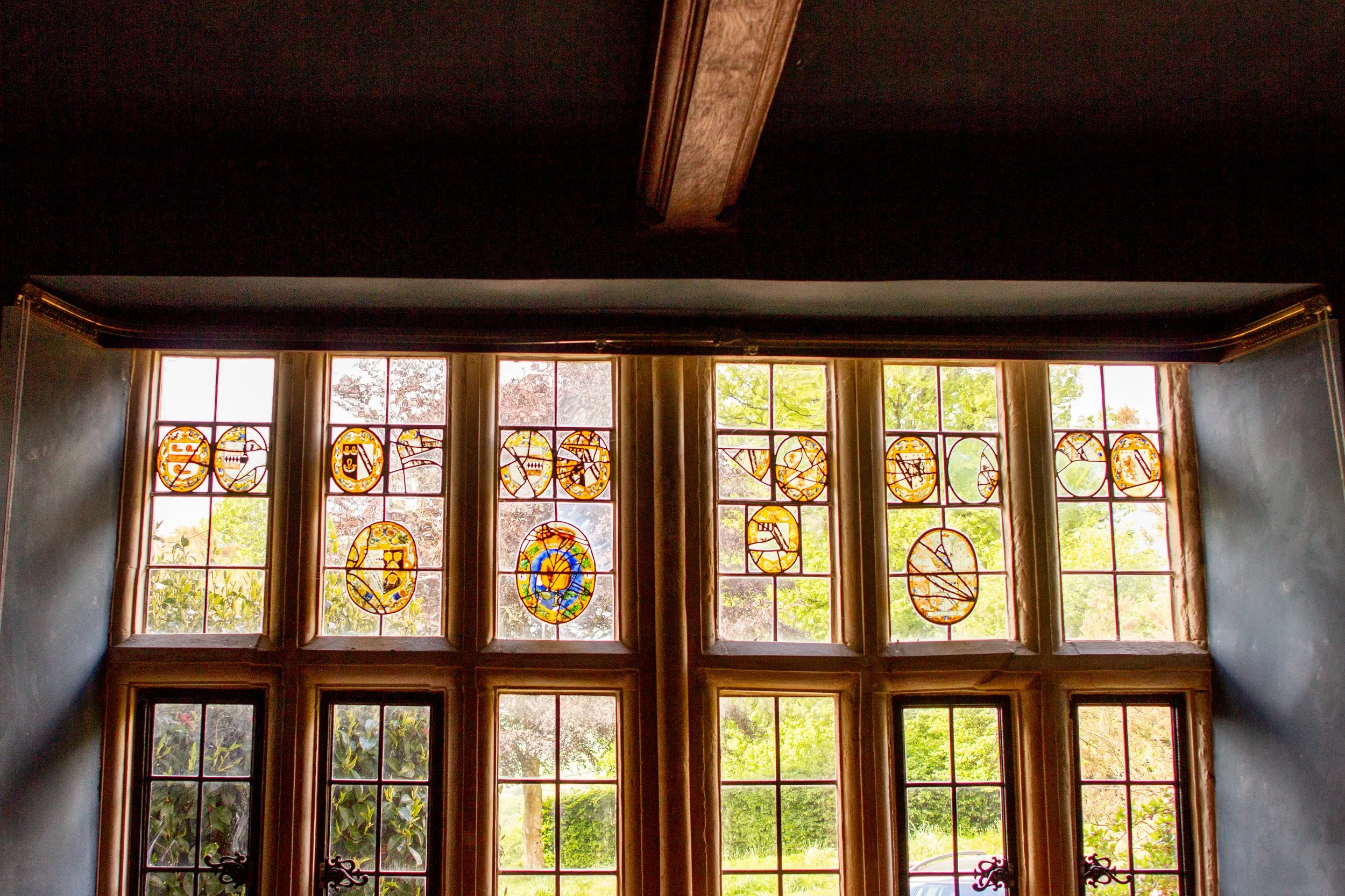 A large window with multiple stained glass panels above a door, showing trees and foliage outside, with sunlight streaming in.