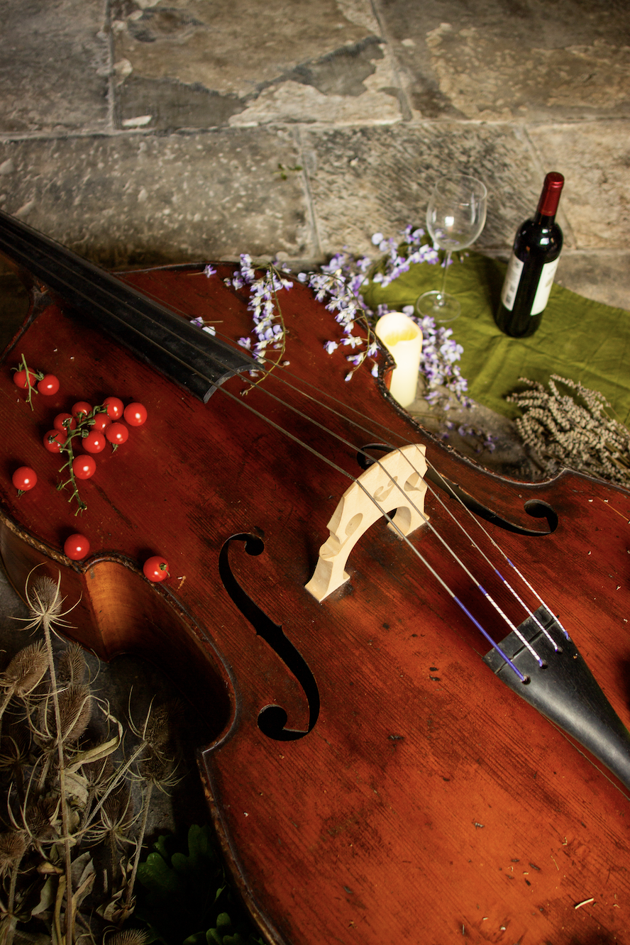 A vintage violin with a bow on top, decorated with red berries and purple flowers, set against a stone wall. A wine bottle, a wine glass, and a candle are on a green cloth nearby.