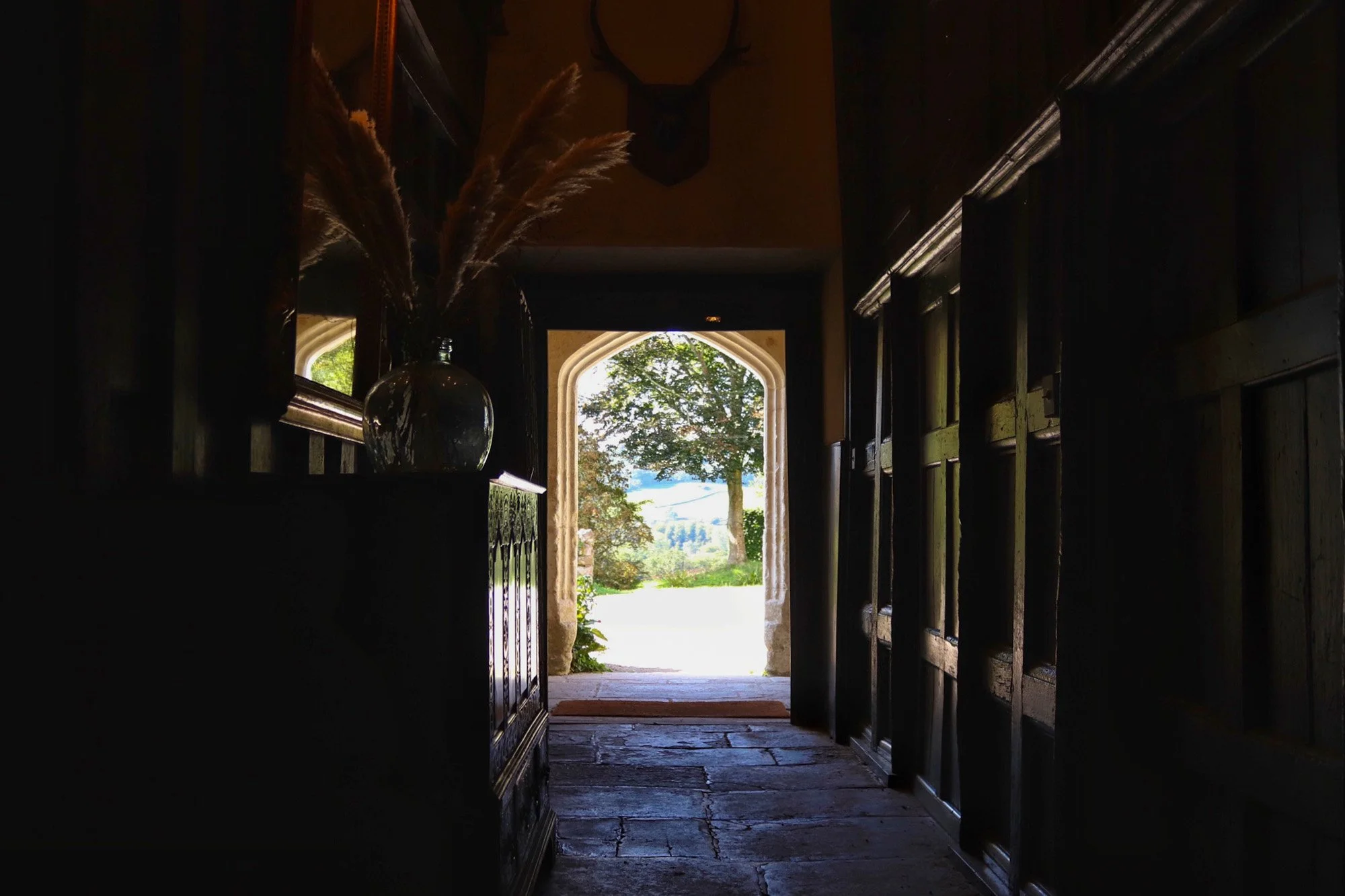 Dark interior corridor leading to a bright outdoor scene with trees and a blue sky visible through an arched doorway.