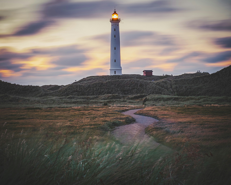 A white lighthouse on top of a small hill - in the evening. Light is on.