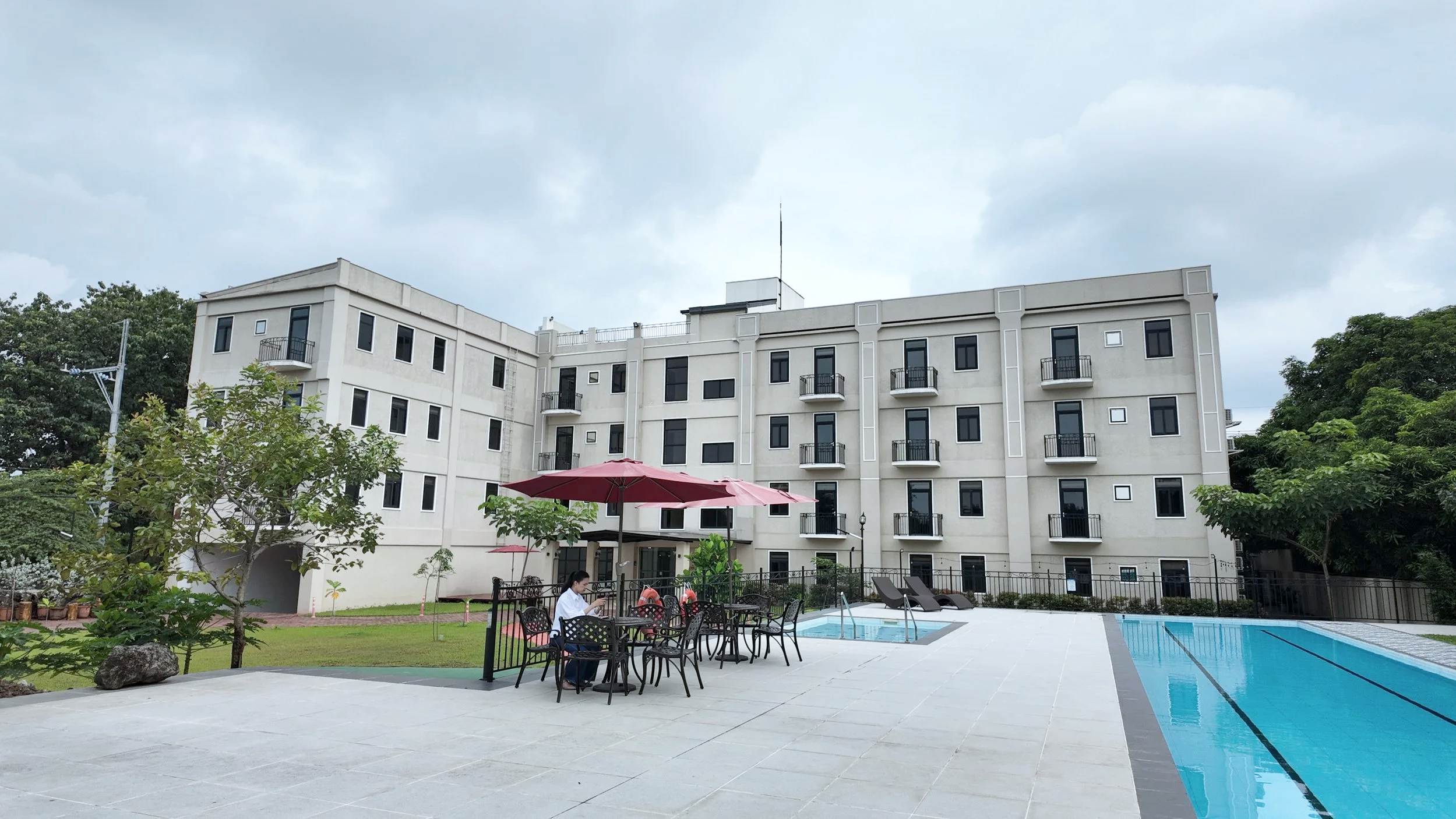Outdoor swimming pool area with patio tables, chairs, and umbrellas in front of a modern white apartment building with multiple balconies, surrounded by greenery and a cloudy sky.