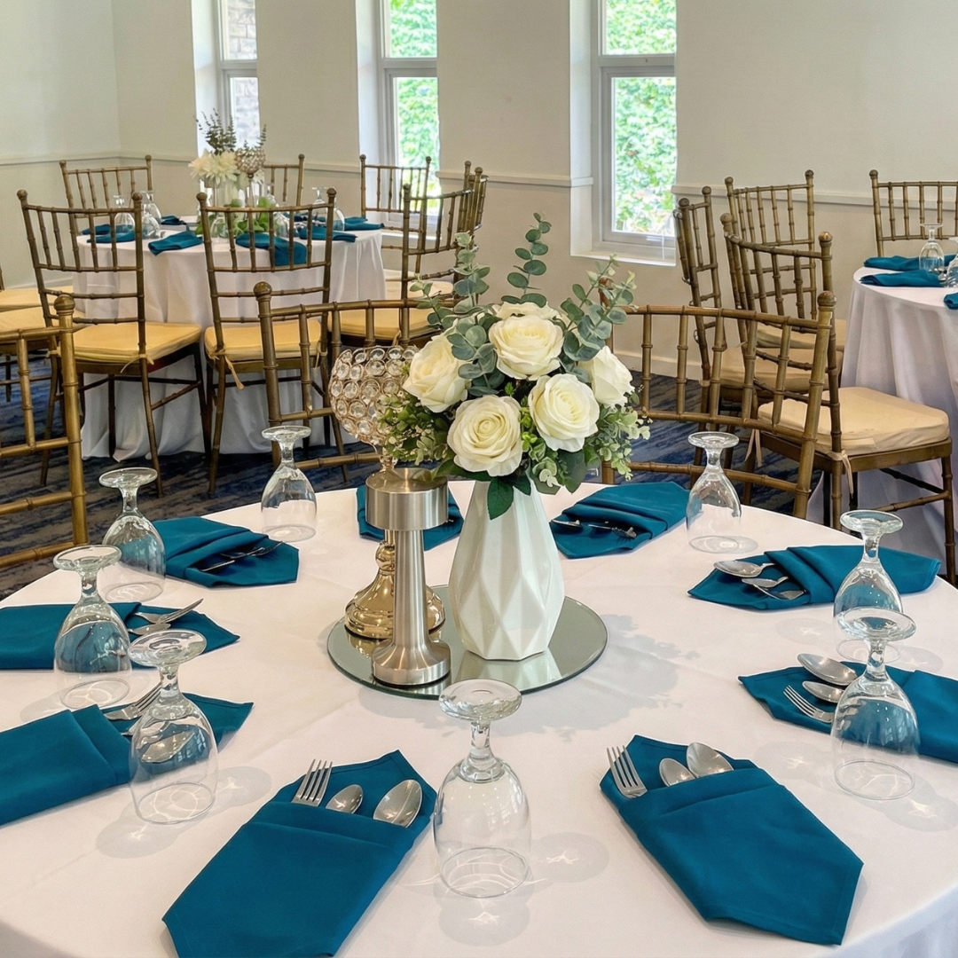 Elegant round table set for a formal event with white tablecloth, blue napkins, silverware, upside-down wine glasses, and a floral centerpiece of white roses and greenery in a white vase. Gold Chiavari chairs and additional tables with similar setups are in the background, with windows and lush greenery outside.