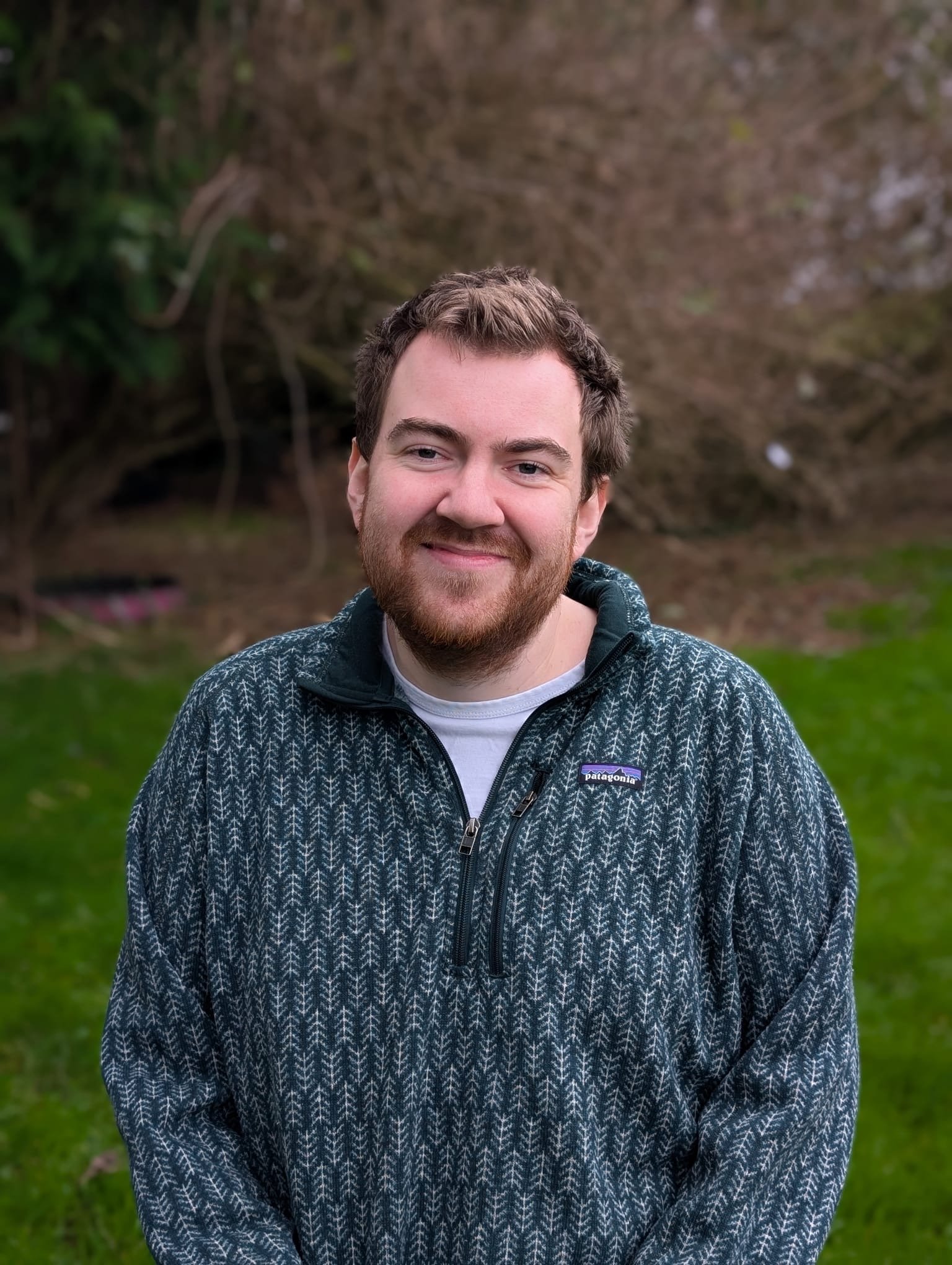 A young man with brown hair and beard smiling outdoors in front of trees and grass, wearing a patterned Patagonia fleece jacket.