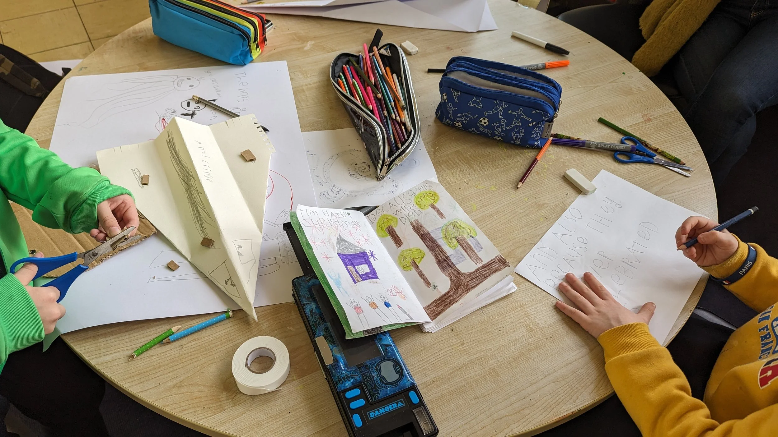 Children's hands engaged in arts and crafts activities on a round table, surrounded by colored pencils, scissors, sketchbooks, and drawings.