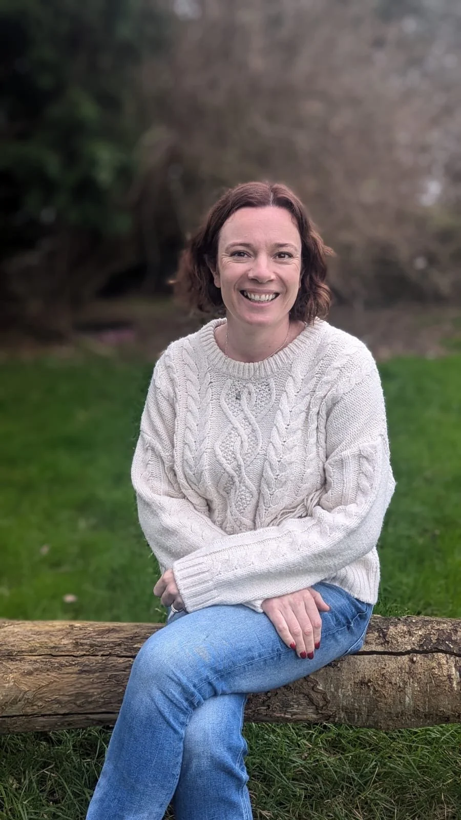 A woman with short brown hair sitting on a wooden log outdoors, smiling, wearing a cream knitted sweater and blue jeans, with a grassy area and trees in the background.