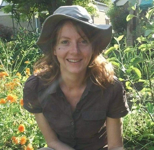 A woman with short red hair smiling, wearing a brown turtleneck sweater, standing outdoors among green foliage.