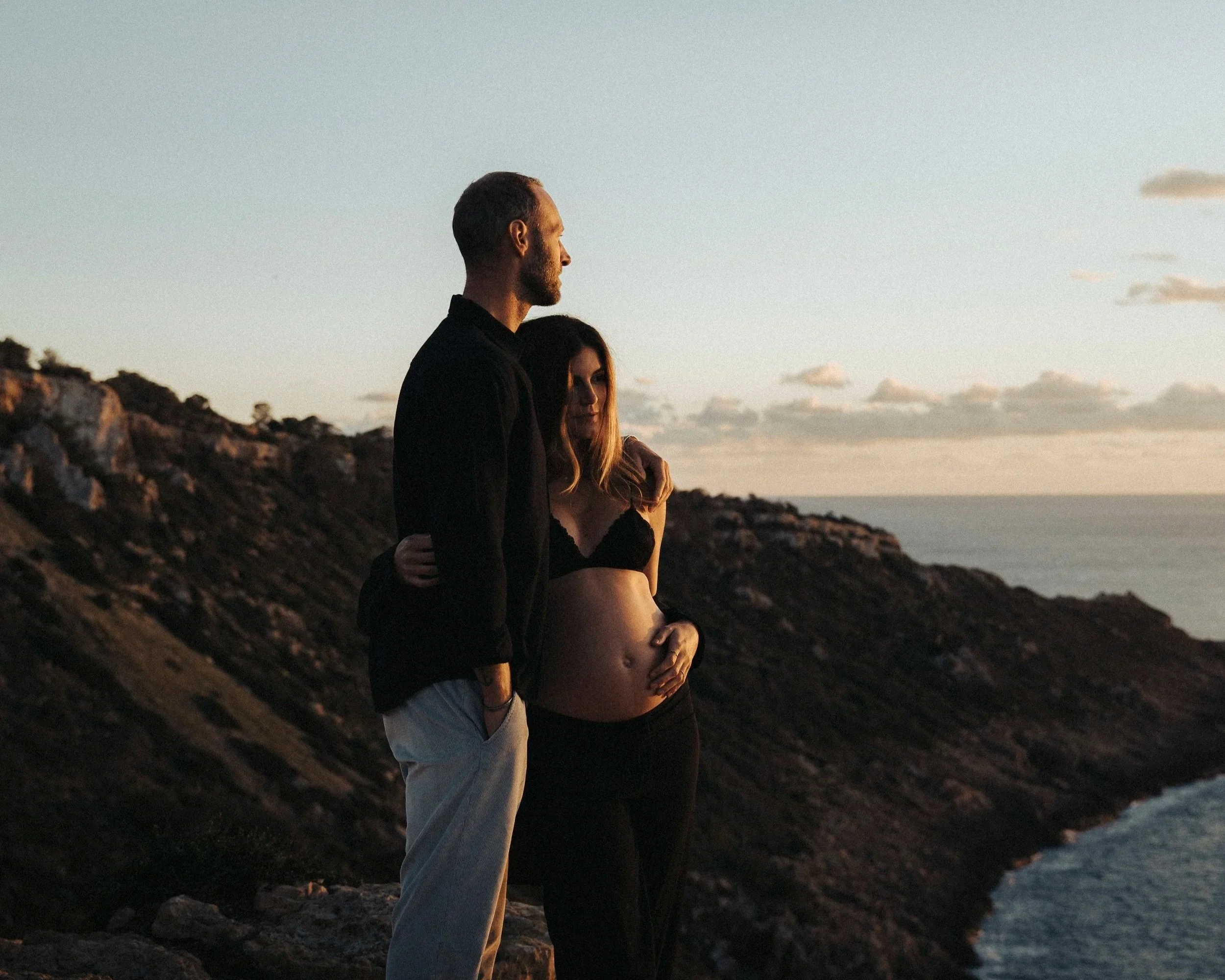 A man and woman stand close together on a rocky coast at sunset, with the man holding the woman by her shoulders. The woman is pregnant, wearing a black bra and black pants, with her hand resting on her baby bump. The man is wearing a black jacket and beige pants. The sky is partly cloudy and the ocean is visible in the background.