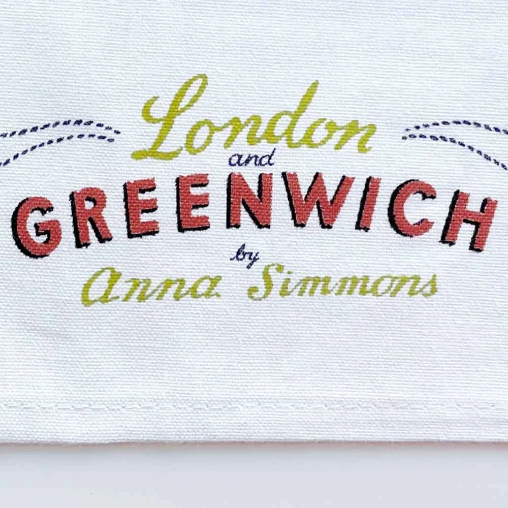 Photograph of the edge of a white tea towel featuring yellow, orange and black hand lettered “London and Greenwich by Anna Simmons”, illustrated by Anna Simmons