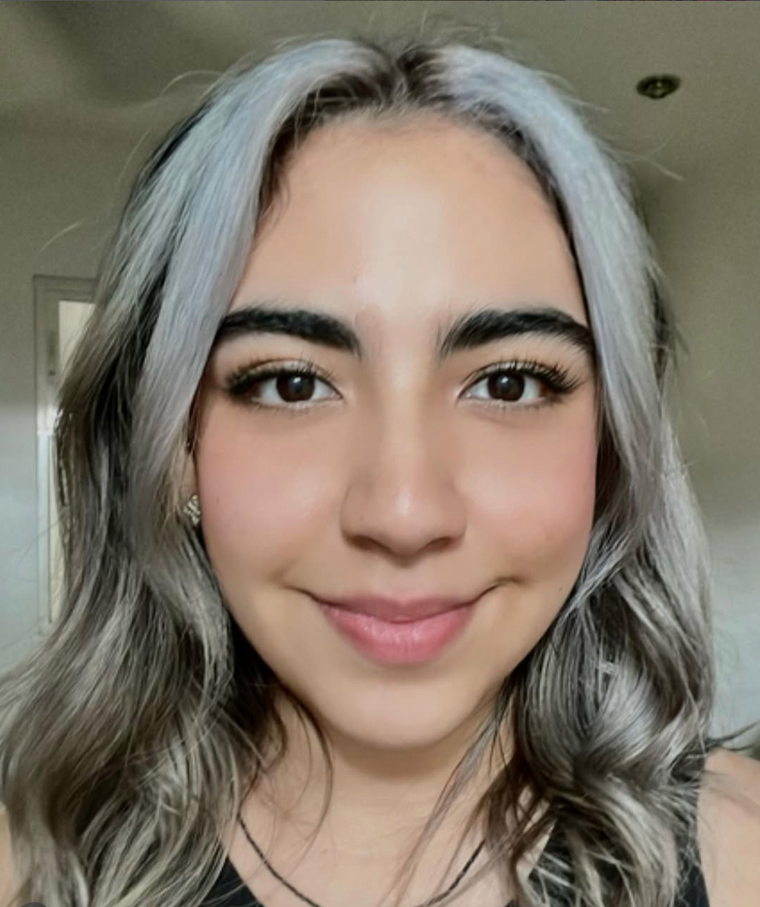 A young woman with silver hair styled in loose curls, dark eyebrows, and brown eyes, smiling in an indoor setting.