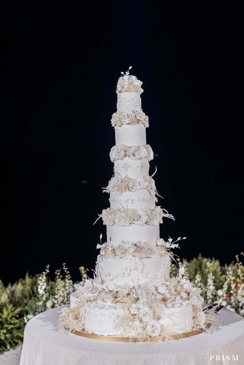 Tall white wedding cake with multiple tiers decorated with white roses and floral accents, placed on a round table with a white tablecloth and floral arrangements in the background, against a dark night sky. Luxury Wedding Planner for Mediterranean