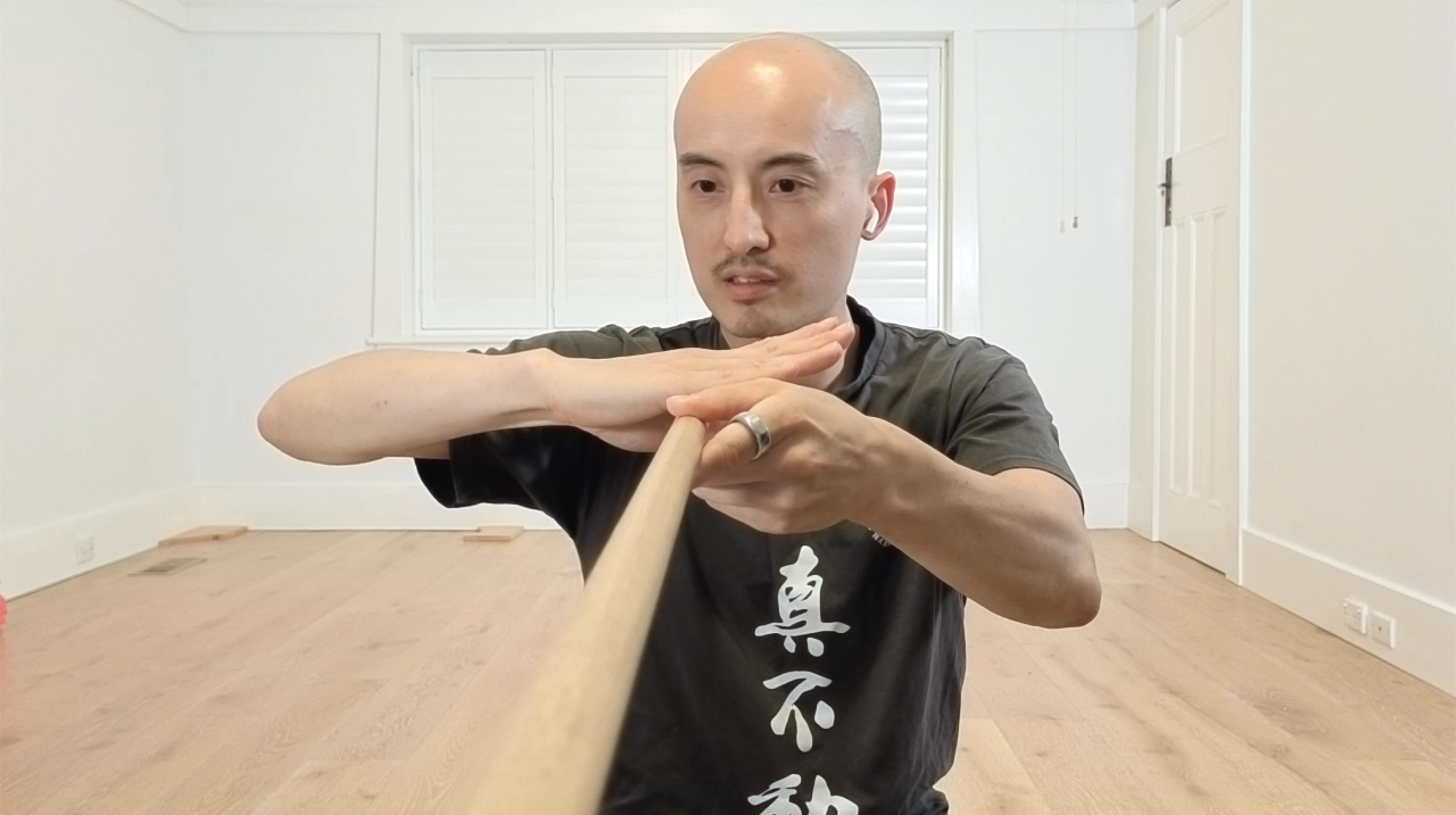 A man practicing martial arts, holding a wooden staff in a home studio with white walls and wooden floor.