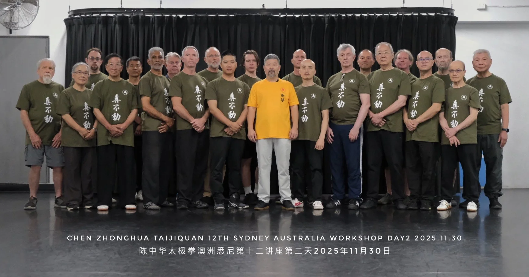 Group photo of 19 individuals in martial arts uniforms standing on a stage with black curtains behind them. One person in the center wears a yellow shirt. Text at the bottom states this was taken during the Chen Zhonghua Taiji Quan 12th Sydney Australia Workshop on November 30, 2025.