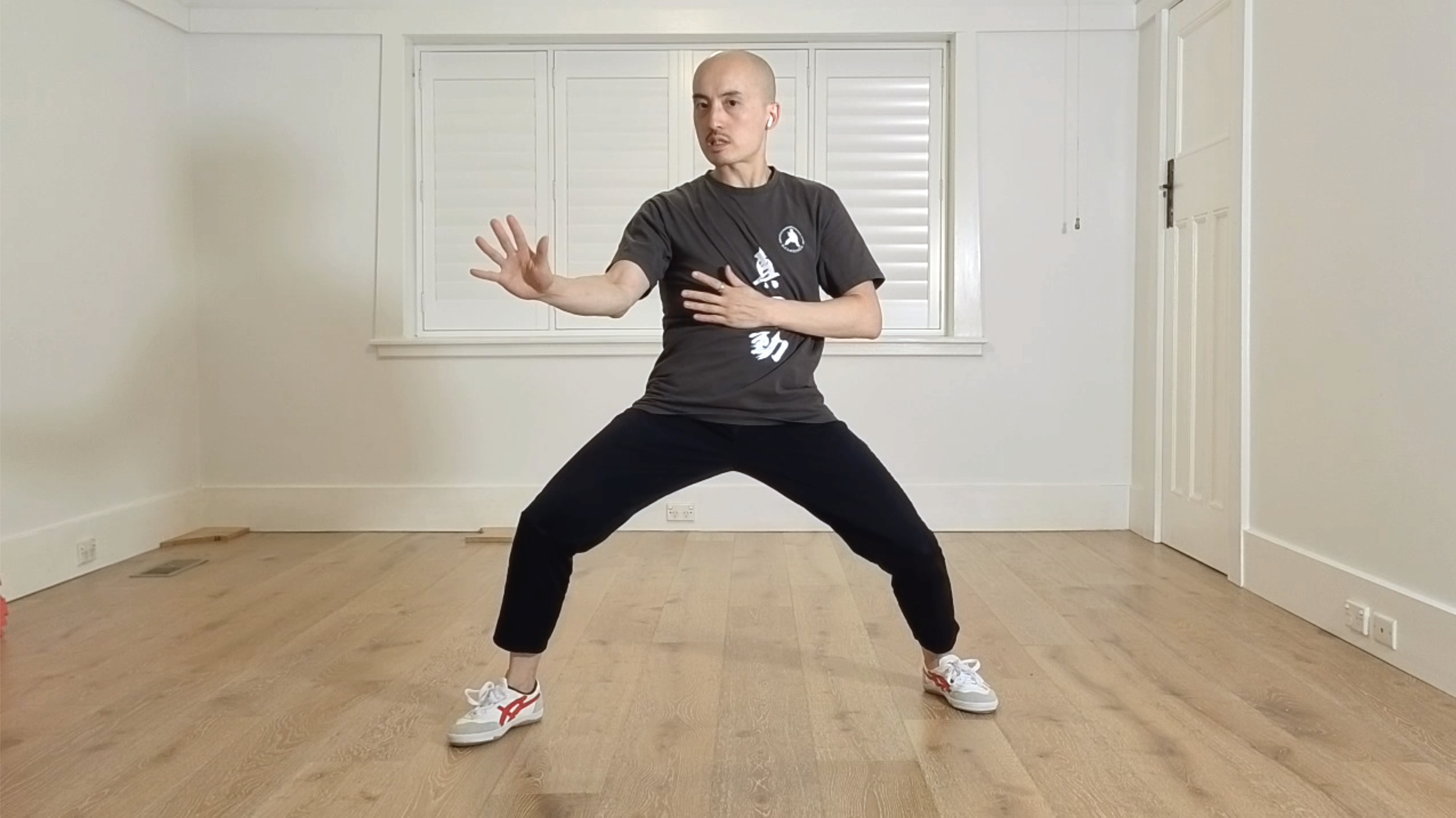 A person practicing a martial arts or tai chi pose in a room with wooden flooring and white walls. The person is wearing a black t-shirt, black pants, white sneakers, and has earbuds in. They are extending their right arm forward with an open hand, and their left arm is crossed over their chest.