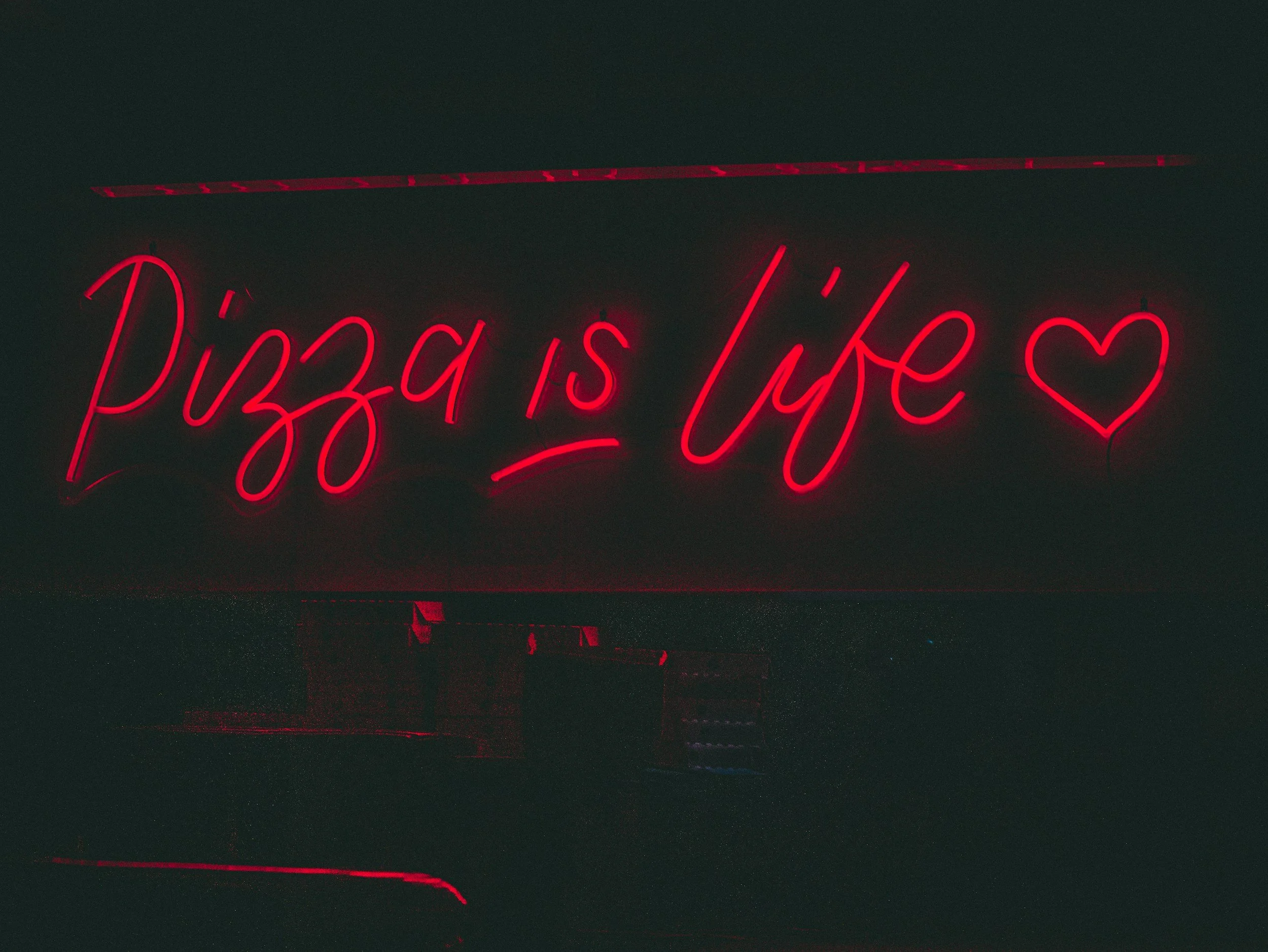 Neon red sign glowing in a dark room, reading 'pizza is life' with a warm, moody ambience.