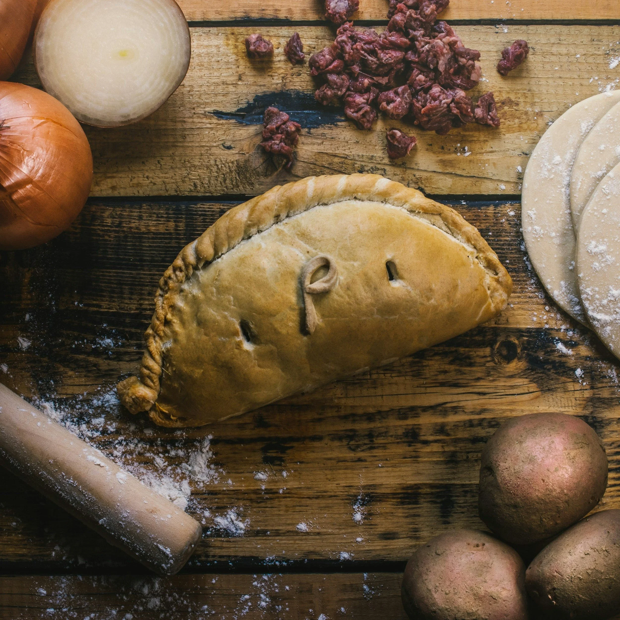 Uncooked Cornish pasty on worktop with key ingredients like flour, pastry, potatoes, steak, and onion nearby.