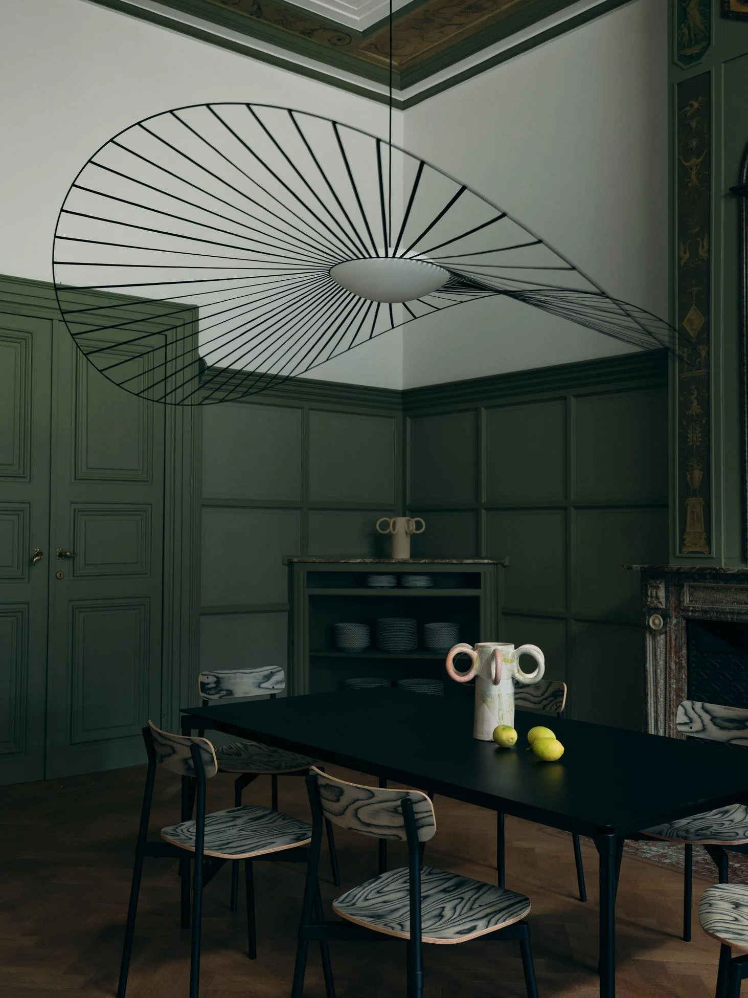 A modern dining room with a dark table, six chairs with wood grain seats, a decorative ceramic pitcher, three lemons, a green paneled wall, and a unique black wire spiral-shaped light fixture on the ceiling.