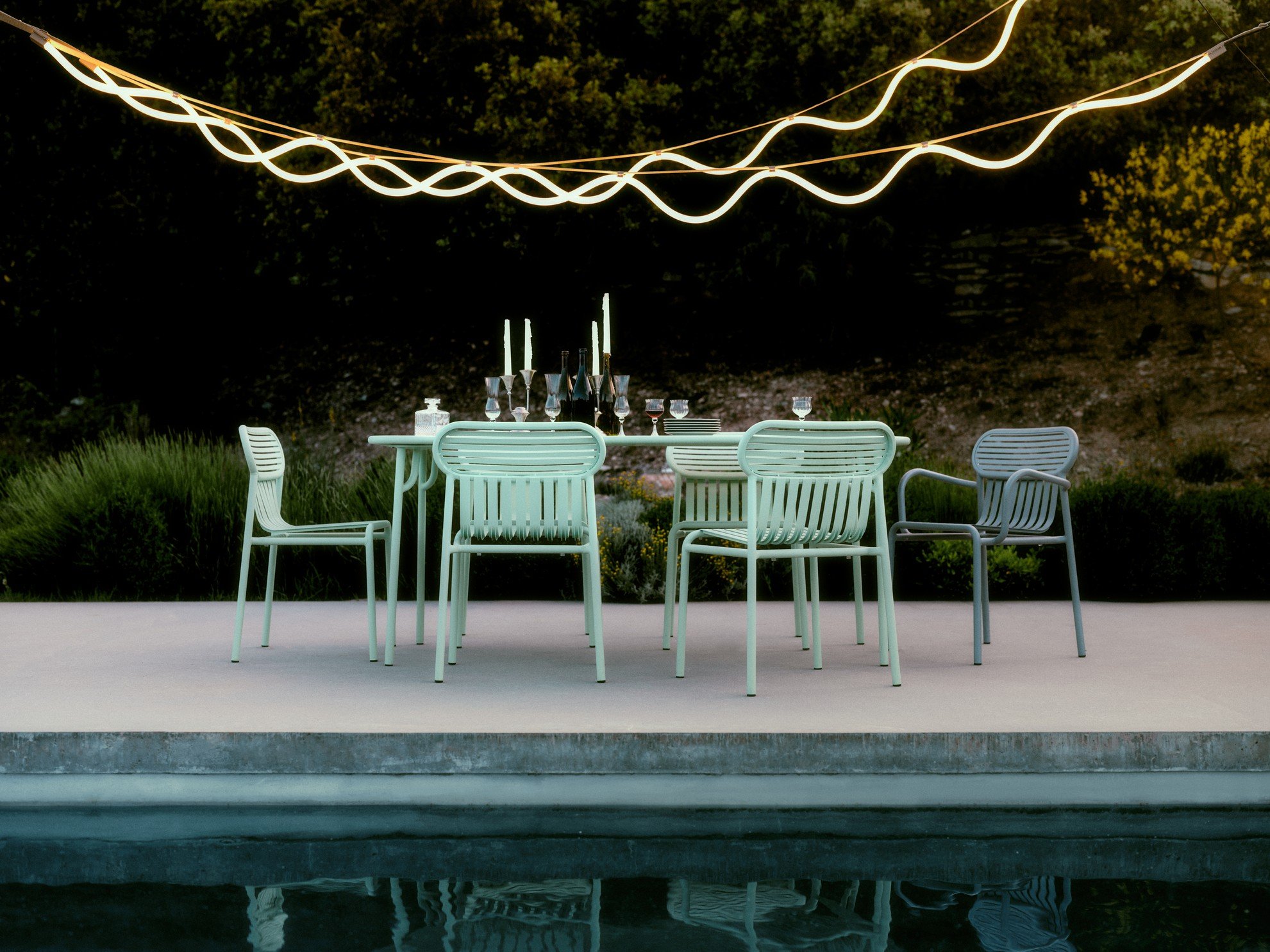 Outdoor dining table set with glasses and candles, surrounded by modern chairs, under decorative string lights, near a pool and lush landscape in the evening.