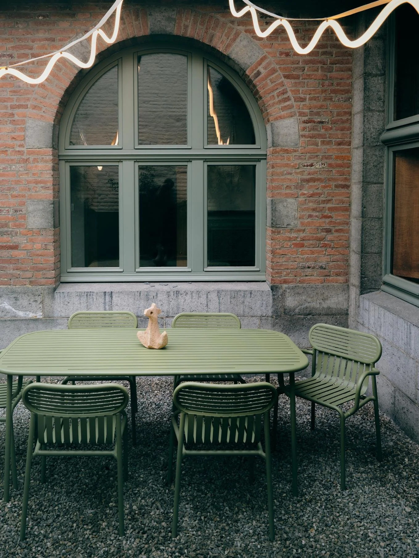 A green outdoor table with six matching chairs around it, and a small decorative llama figure in the center, set on a gravel ground against a brick wall with a large arched window.