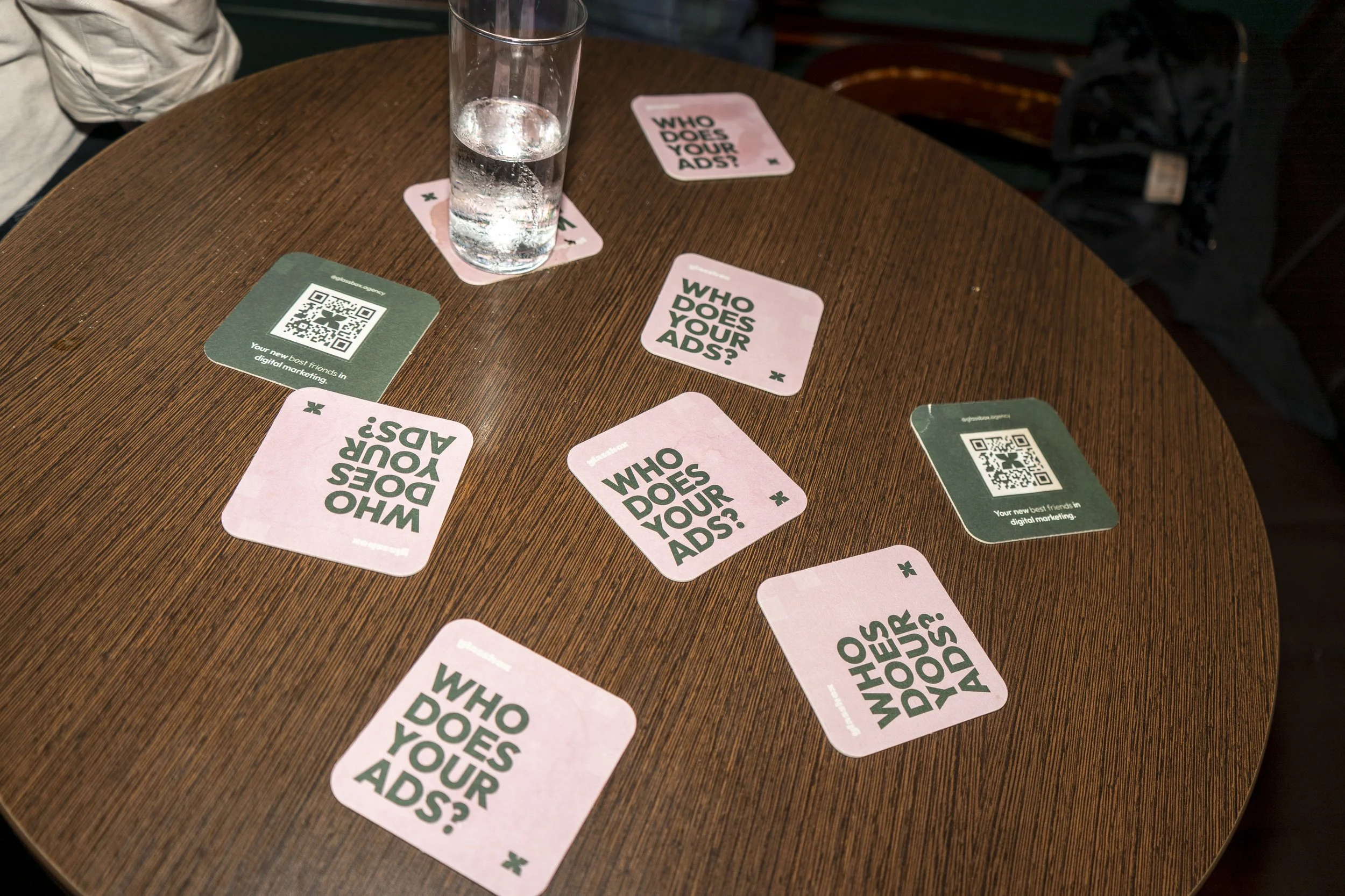 Round wooden table with multiple pink and green cards, a tall glass of water, and a black backpack in the background. The pink cards read 'WHO DOES YOUR ADS?' and the green cards contain QR codes.