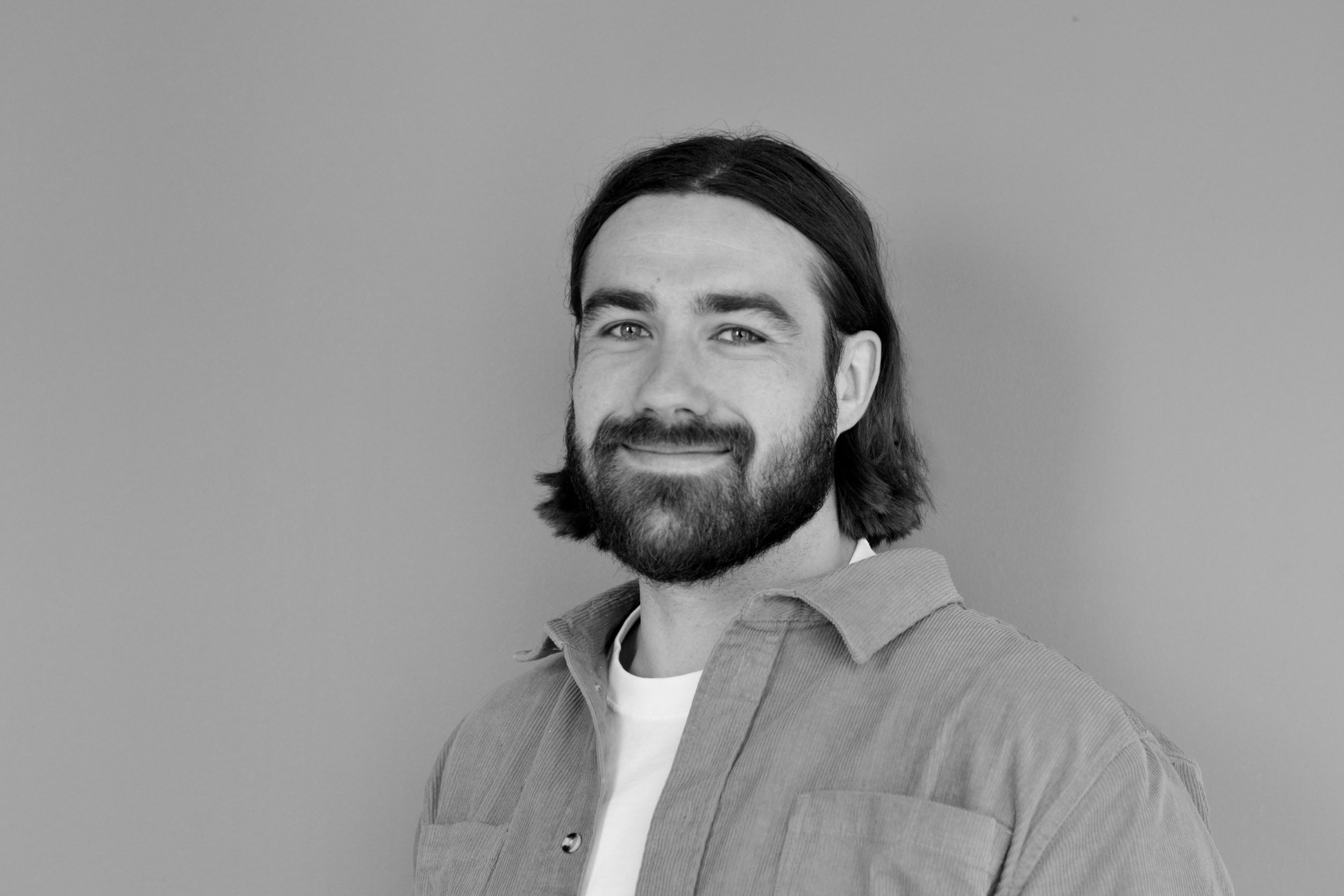 A black and white portrait of a man with long dark hair, beard, and mustache, smiling slightly, wearing a collared shirt over a white t-shirt against a plain background.