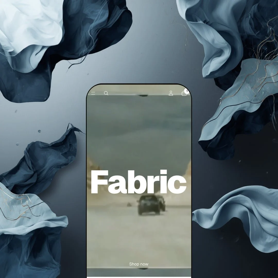 Fabric Shopify theme displayed on a mobile phone showing the video hero section with bold Fabric logo and Shop now button, surrounded by flowing fabric textures in dark blue and grey tones.