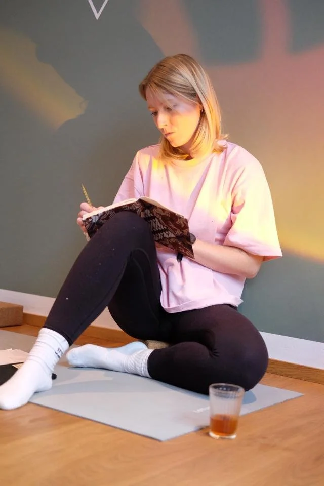 Junge Frau sitzt auf Yoga-Matte auf dem Boden und schreibt in ein Notizbuch, trinkt ein Glas Tee.